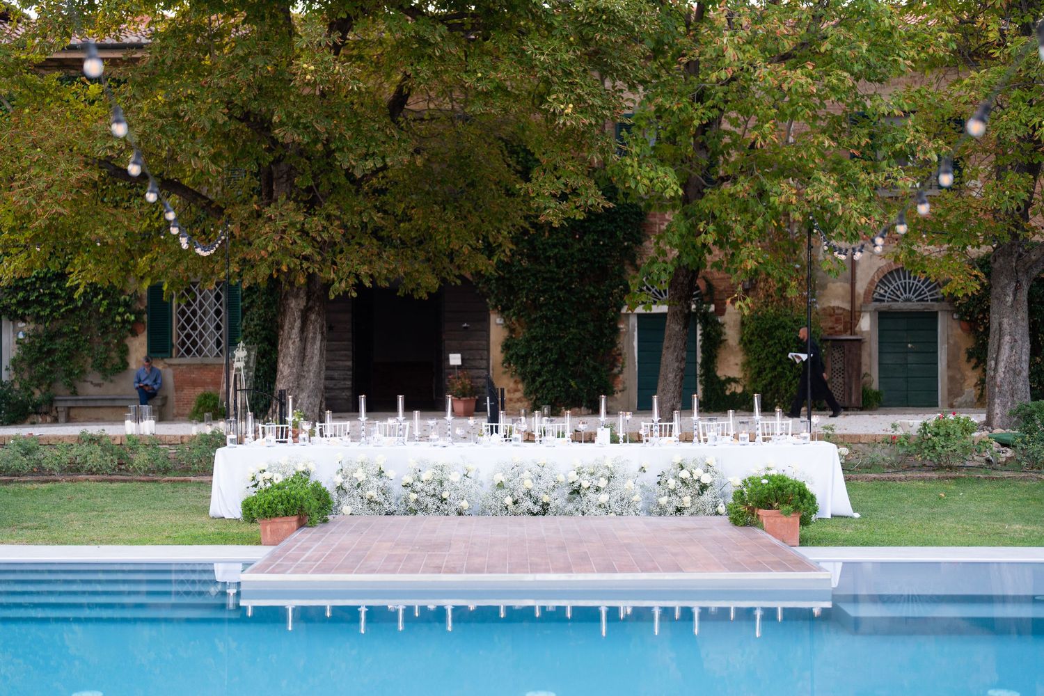 An outdoor wedding reception setup with white tables and chairs arranged near a swimming pool.