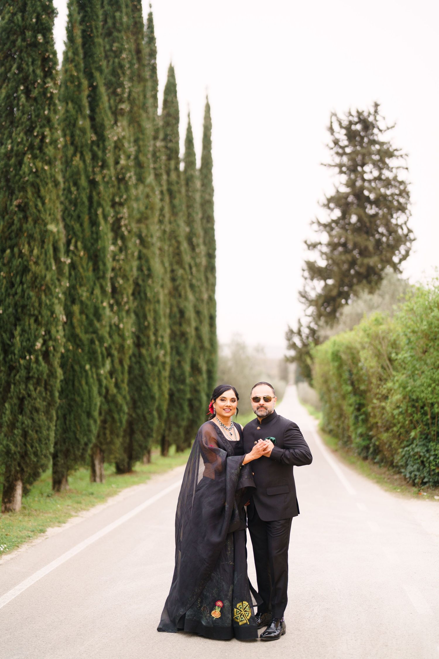 Couple in formal black attire share intimate moment on cypress-lined path.