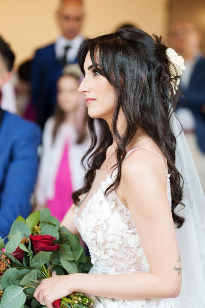 Bride in a lace wedding dress holds a red rose bouquet during an elegant indoor ceremony.