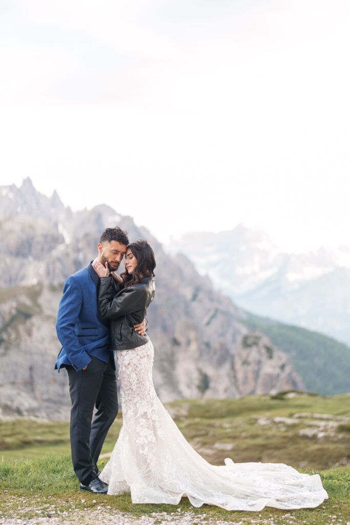 A wedding couple embraces on a mountain meadow with dramatic rocky peaks in the background.