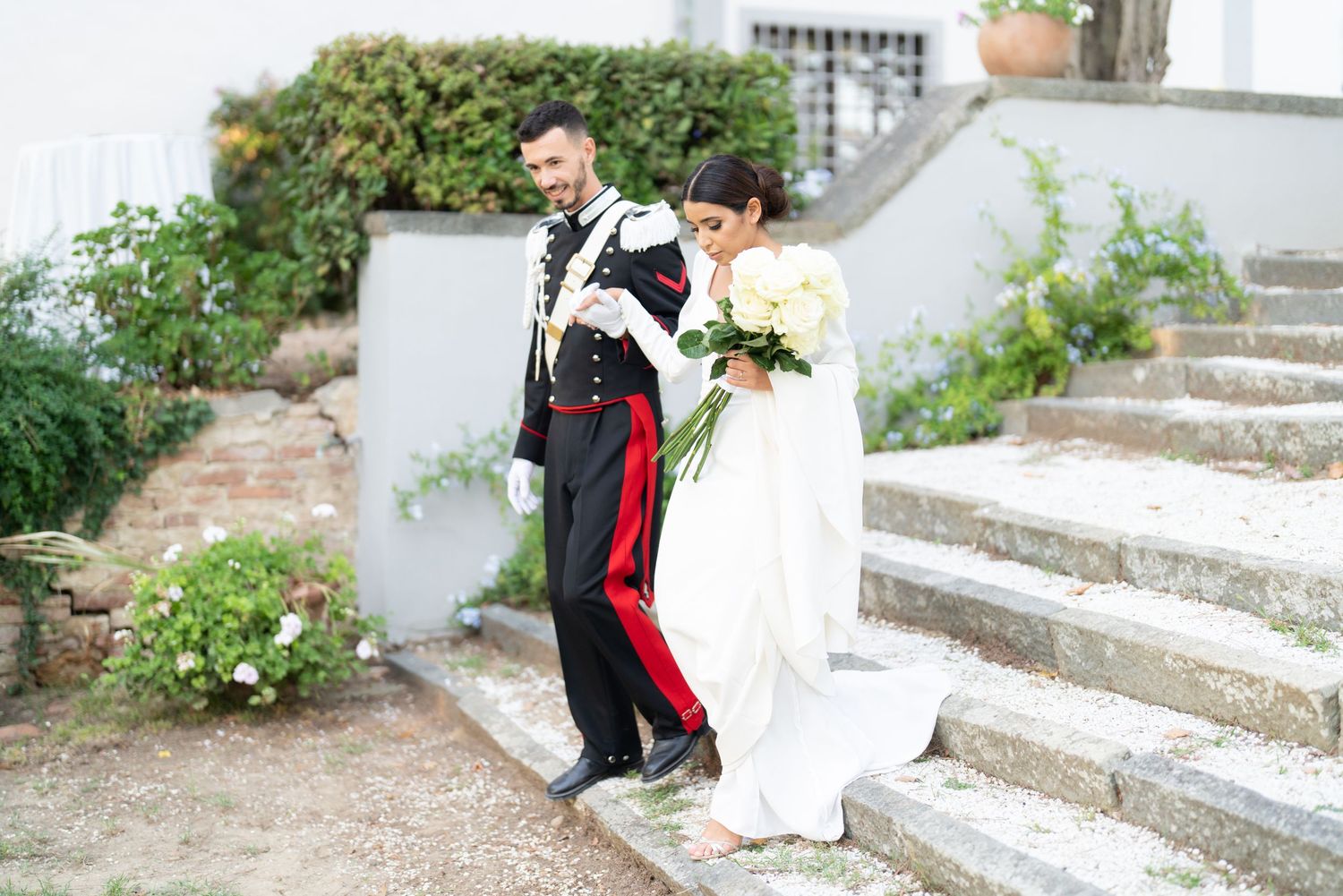 Wedding ceremony participants walk down stone steps in garden setting.