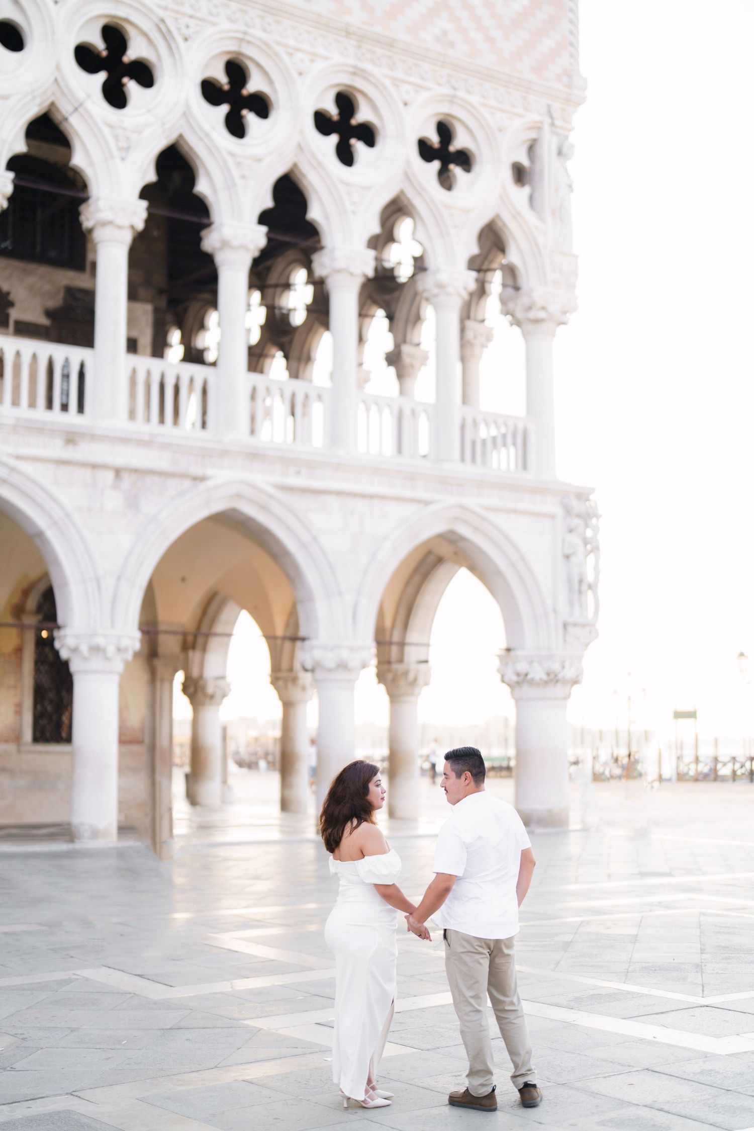 A couple in white outfits shares a romantic moment in front of ornate Venetian Gothic architecture.