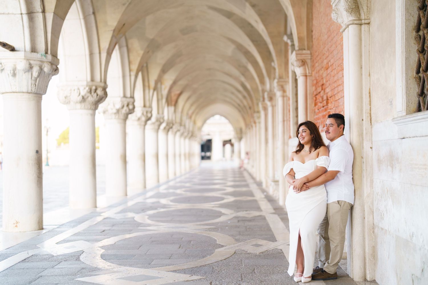 A couple shares a romantic moment in an elegant white stone archway corridor with classical architecture.