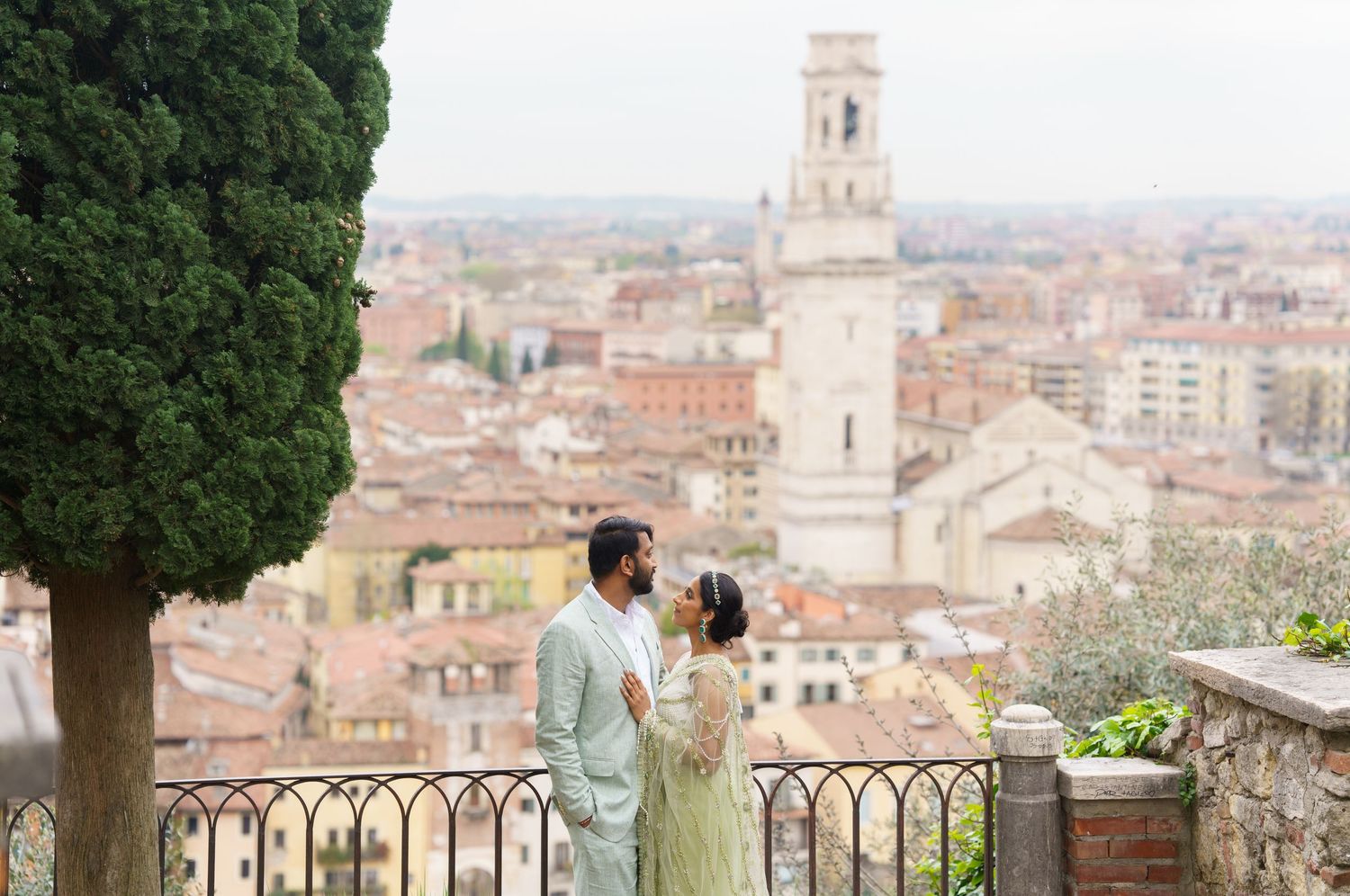 Person admires panoramic cityscape view from hilltop terrace overlooking historic European town with bell tower.