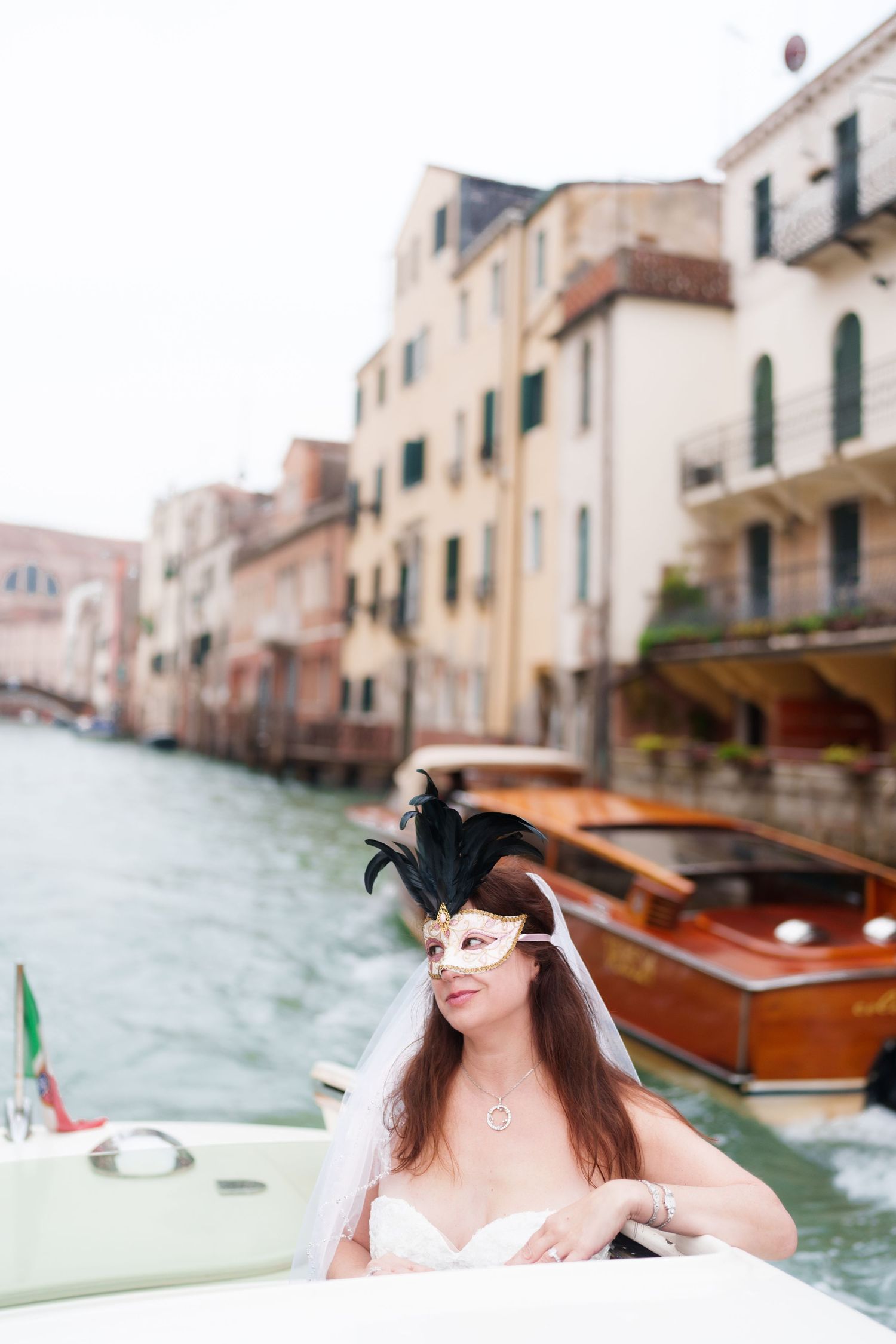 Person wearing a Venetian mask sits in a boat on a canal in Venice with traditional buildings and wooden boats in background.