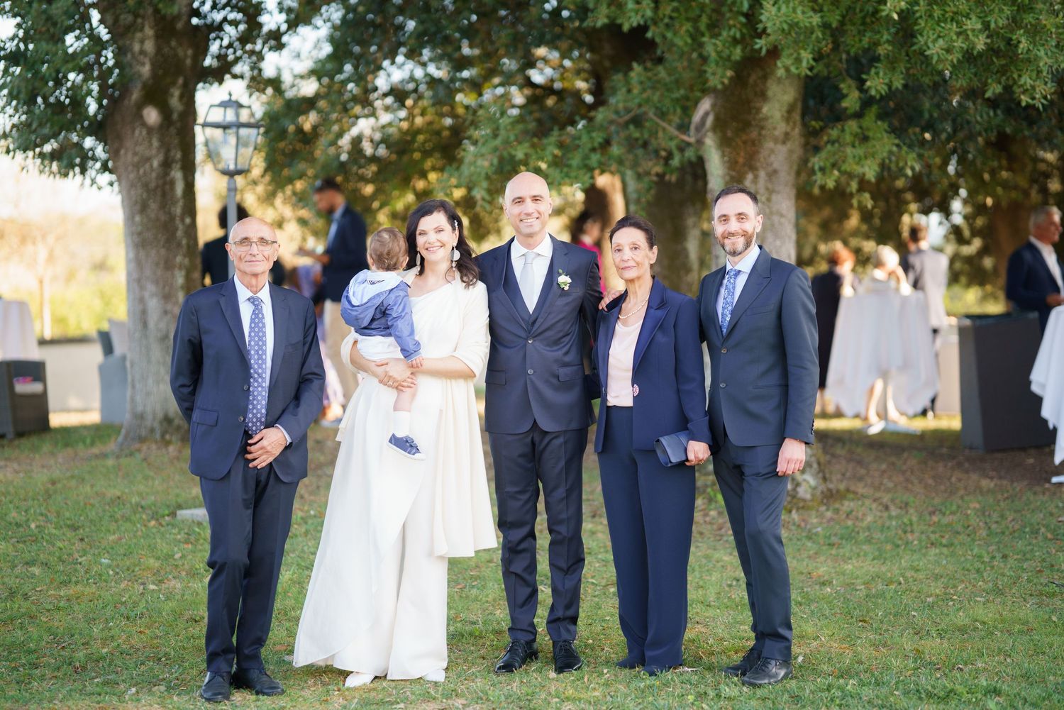 Wedding party members in navy suits and white dresses gathered outdoors on grass.