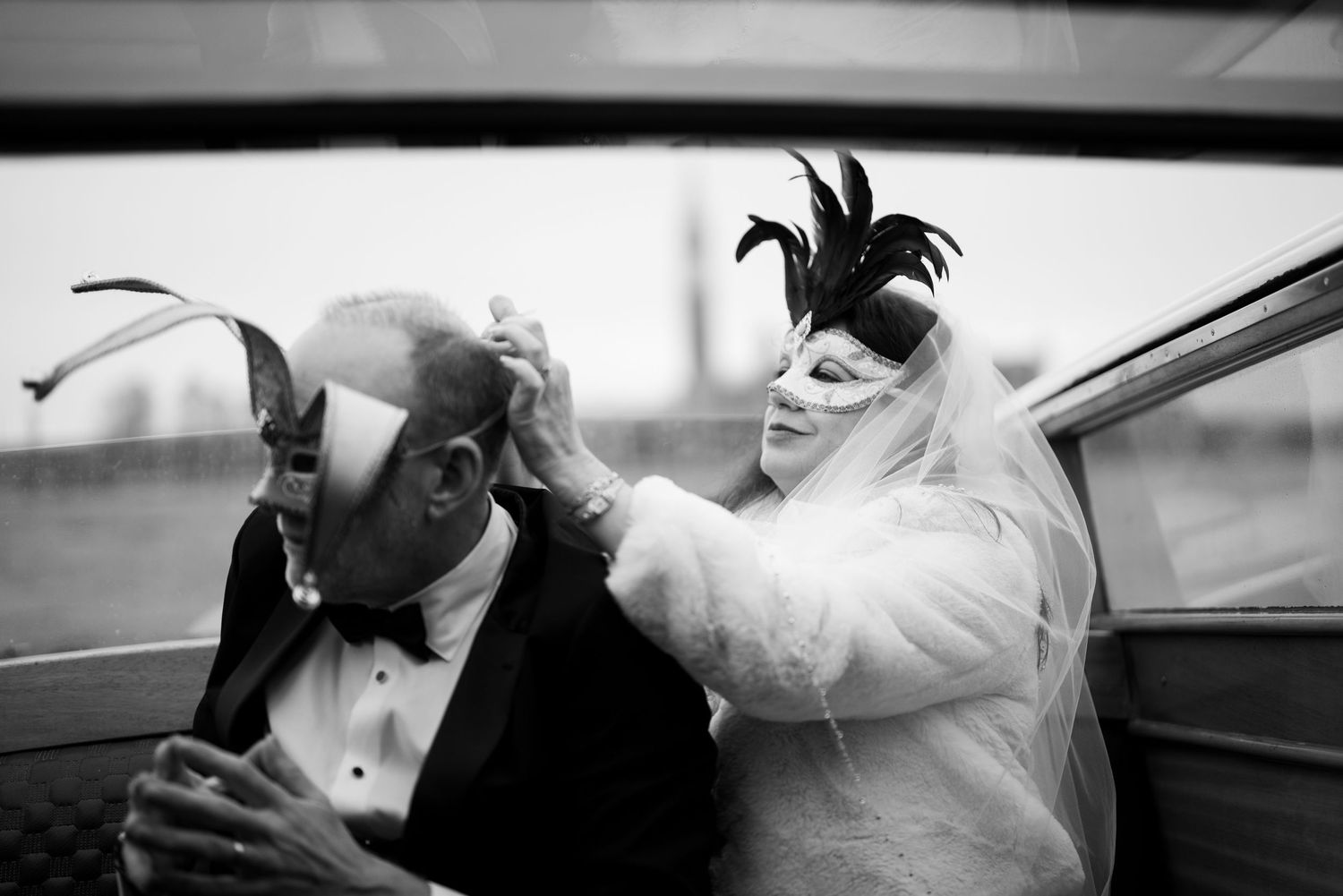 A black and white photo shows two people in masquerade masks and formal attire sitting in a car.