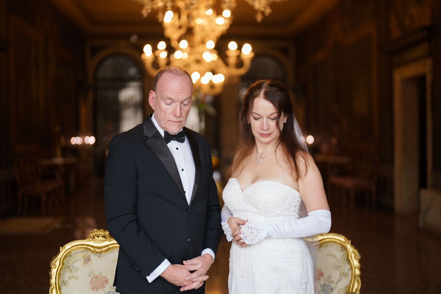Elegant couple in formal wedding attire poses in luxurious ballroom with ornate chandelier and gold furniture.