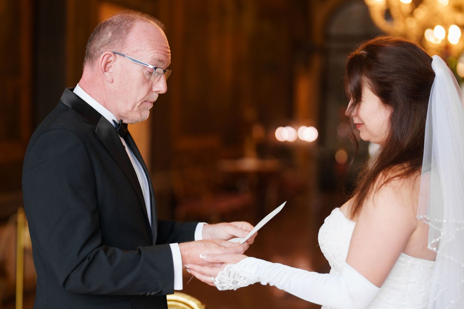 Bride and groom exchange rings during an elegant indoor wedding ceremony with warm lighting and chandeliers.
