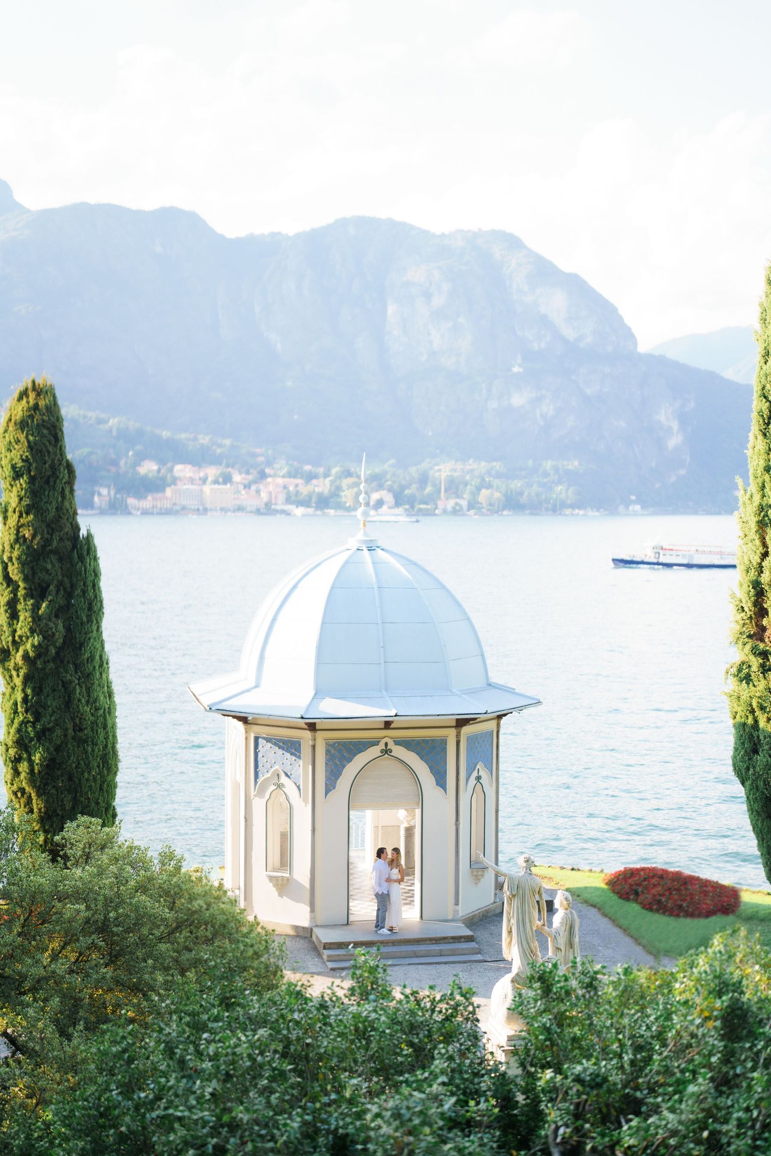 Elegant white gazebos overlook a serene lake with cypress trees and mountains in the background at Lake Como, Italy.