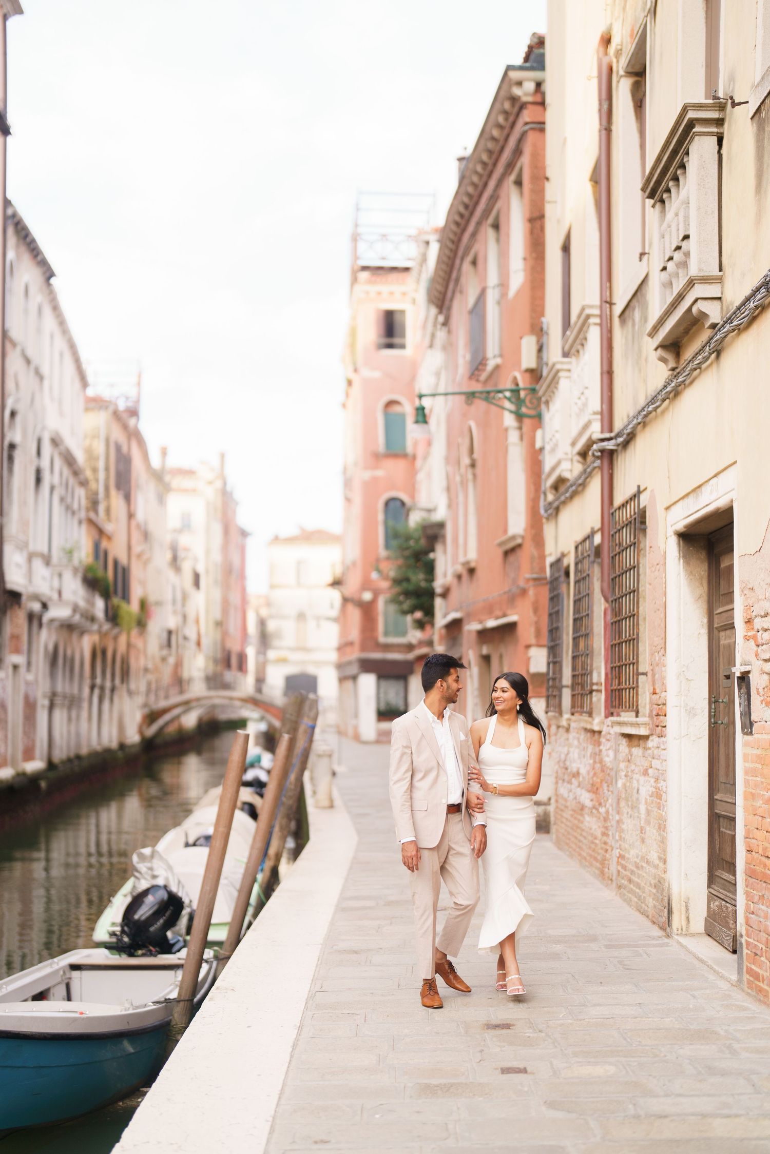 A romantic couple strolls along a scenic canal in Venice, Italy with historic architecture and bridges in the background.