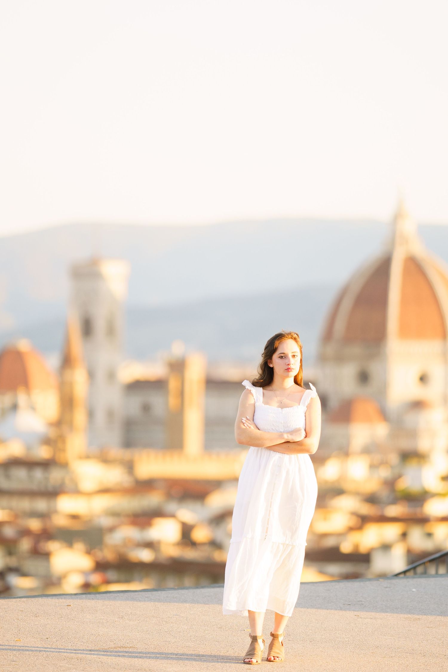 A person in a white dress stands against a golden-hour cityscape of Florence, Italy with the Duomo cathedral visible.