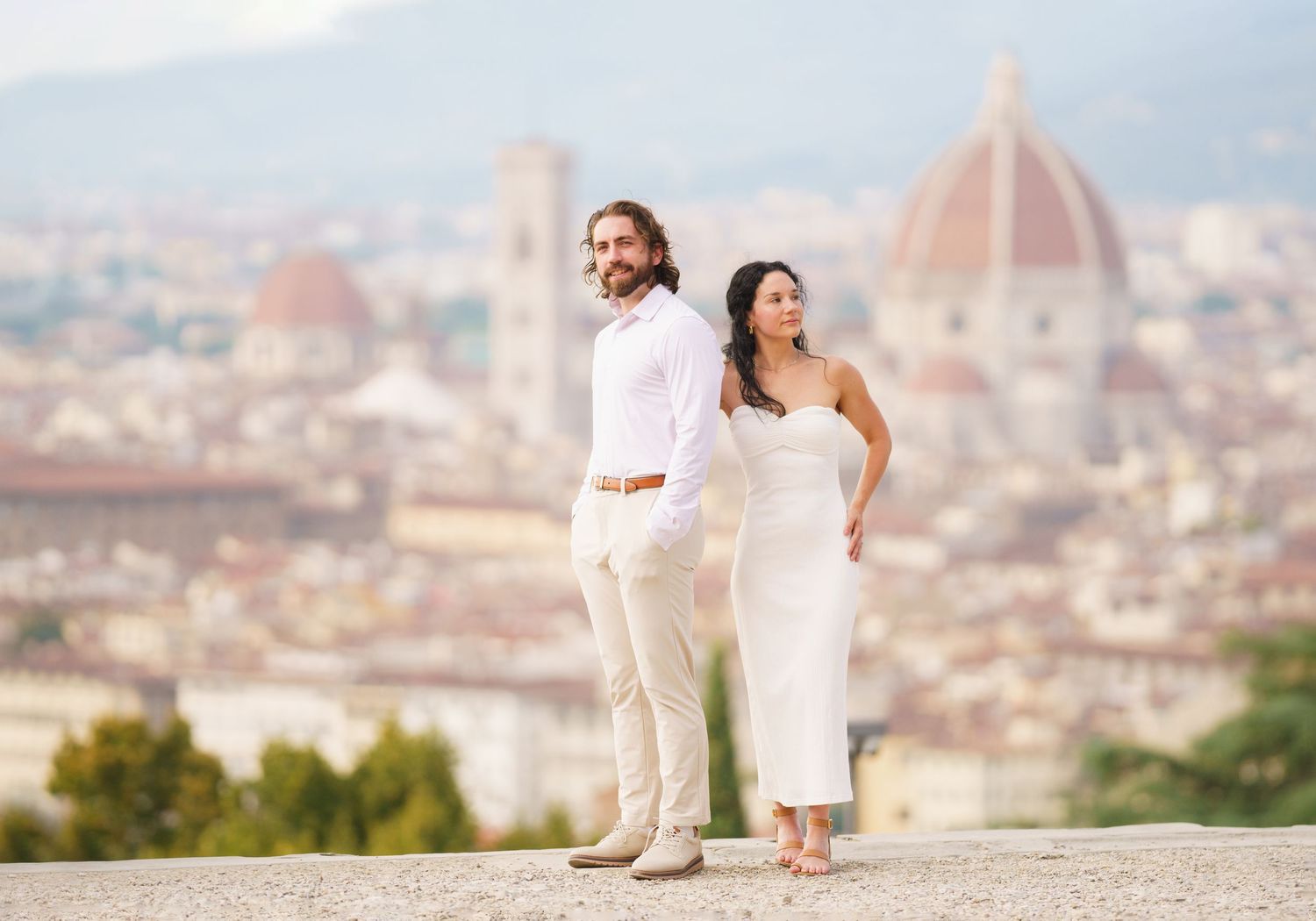 Romantic couple in white outfits walking along Piazzale Michelangelo overlooking Florence's iconic Duomo cathedral.