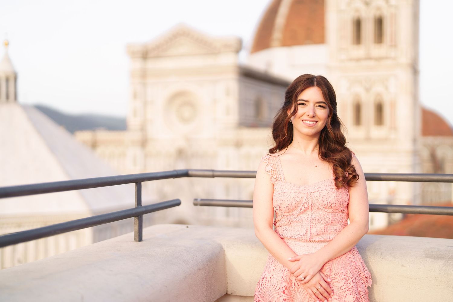 Woman in pink lace dress sits on rooftop terrace with historic cathedral building in background.