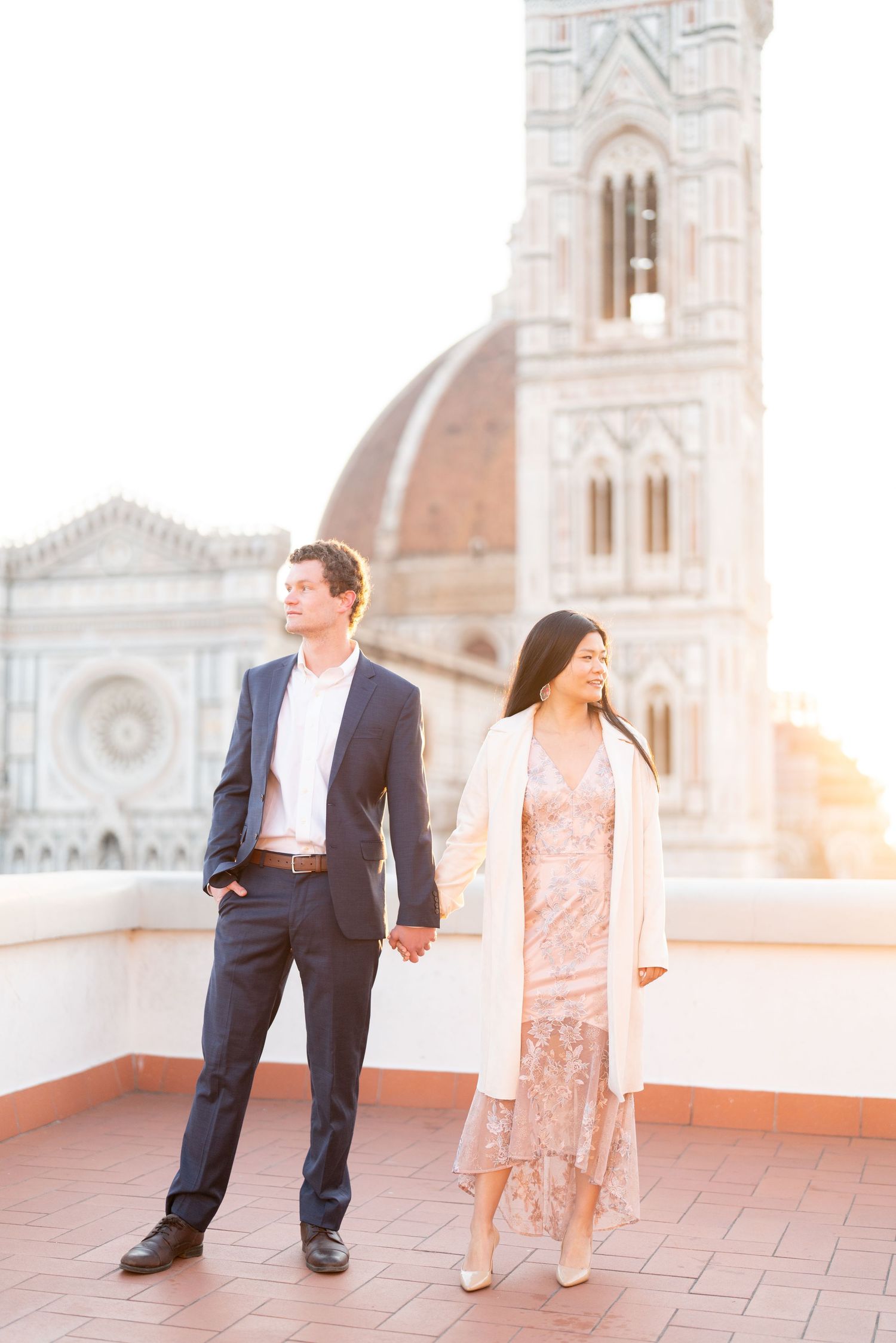A romantic couple walks together on a terrace overlooking Florence's Duomo cathedral during golden hour.