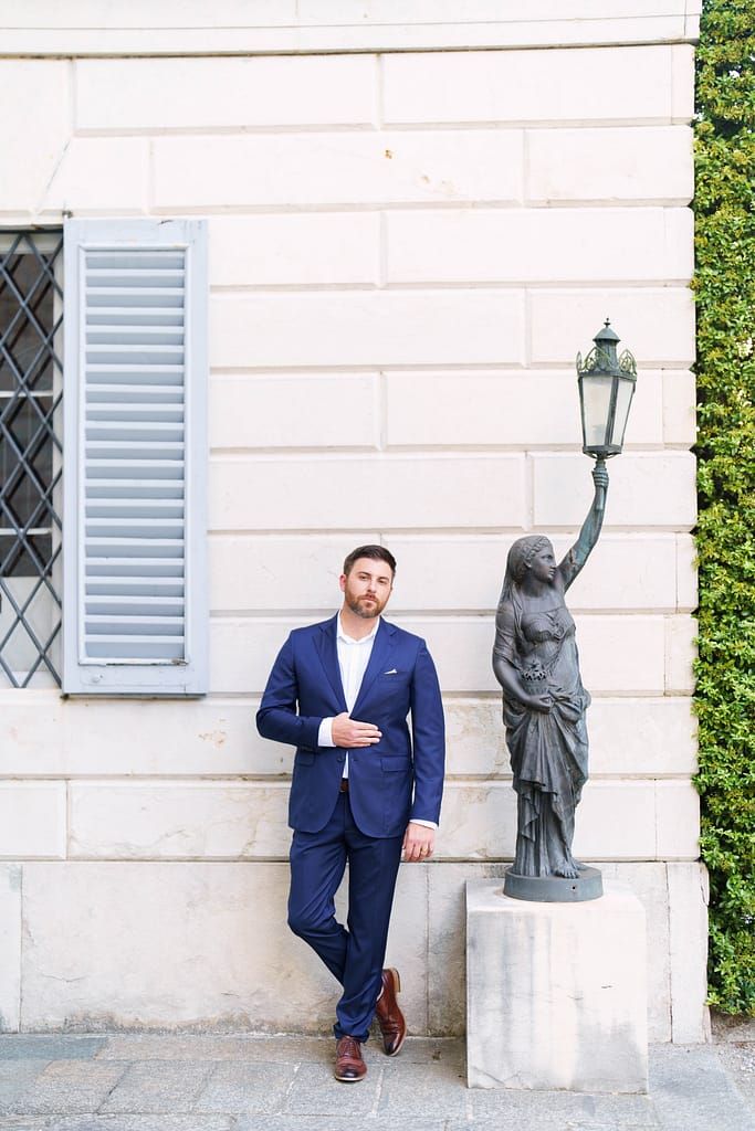 A person in a navy suit poses next to decorative stone statues outside a white building with shuttered windows.