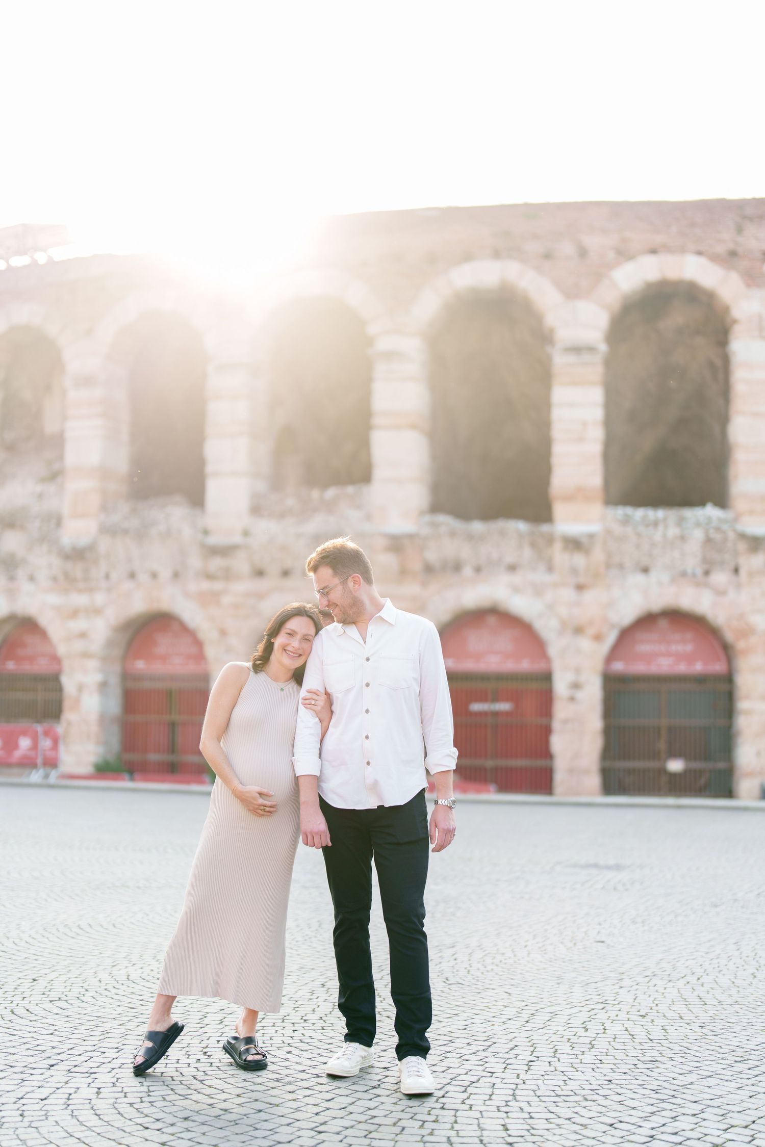 A romantic couple strolls together under stone arches at sunset in a European plaza with cobblestone pavement.