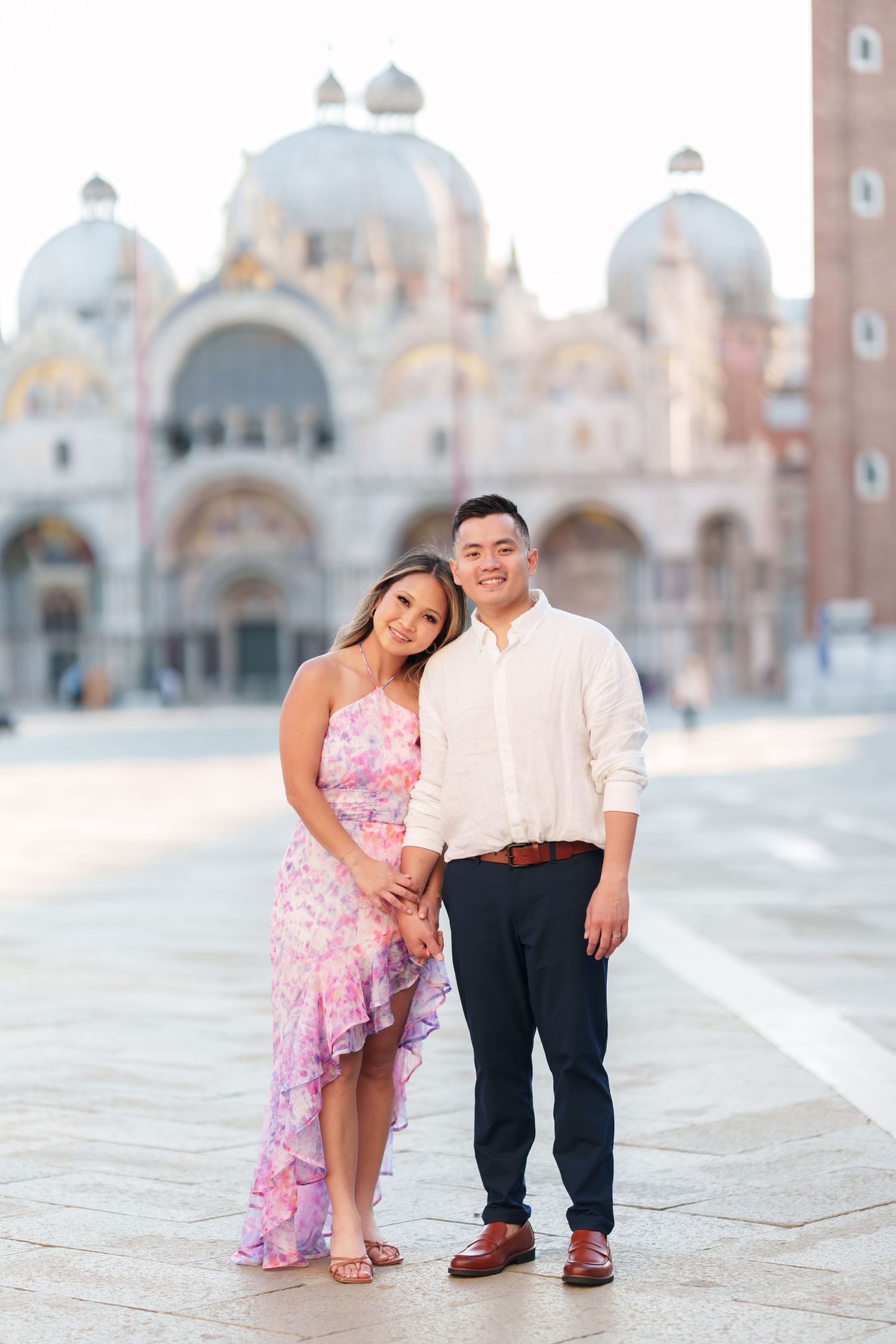 Couple posing in St. Mark's Square, Venice, woman in floral dress, man in white shirt, Basilica in background.