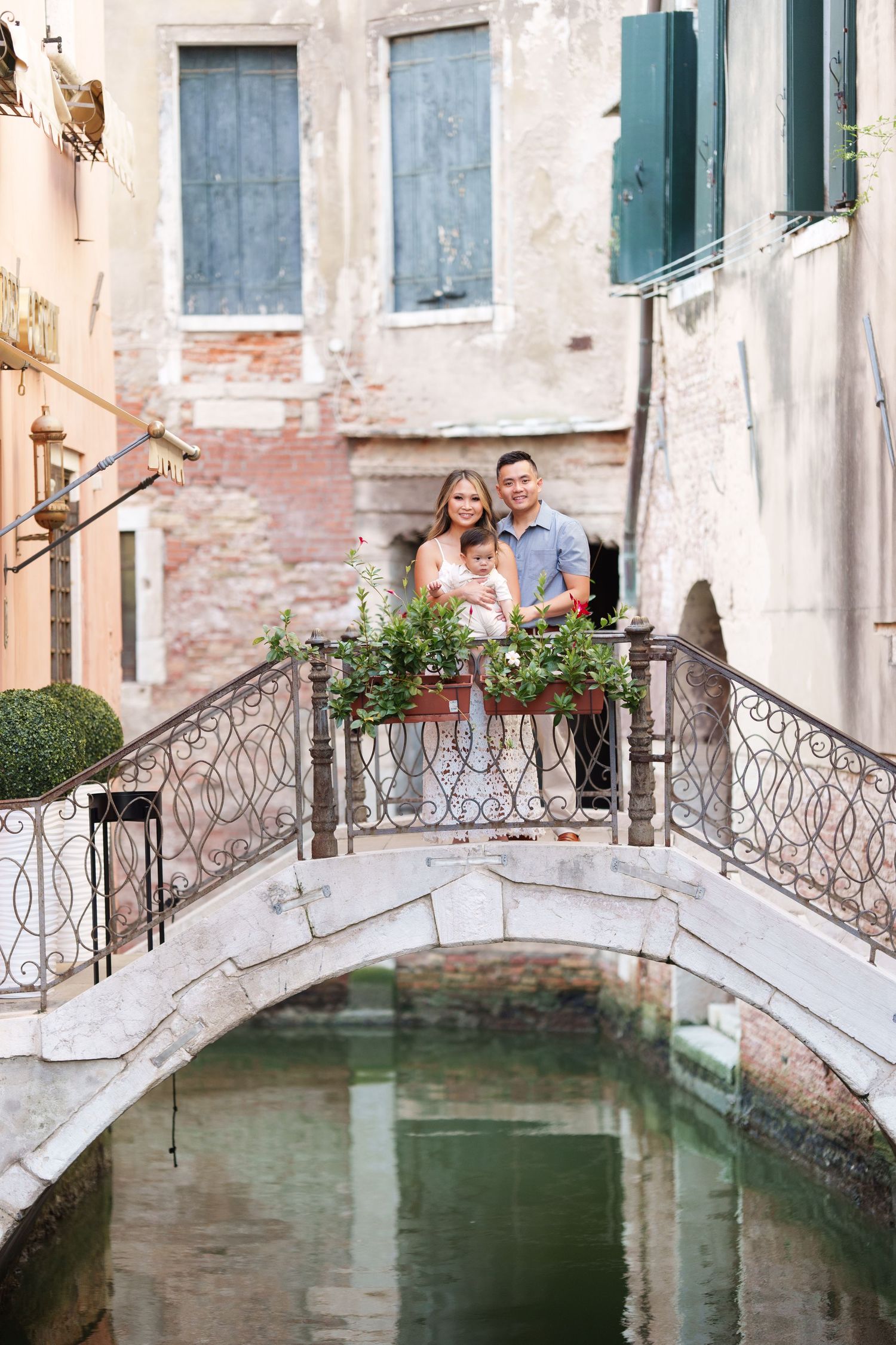Family of three posing on a charming stone bridge over a Venice canal, surrounded by colorful flowers and historic buildings.