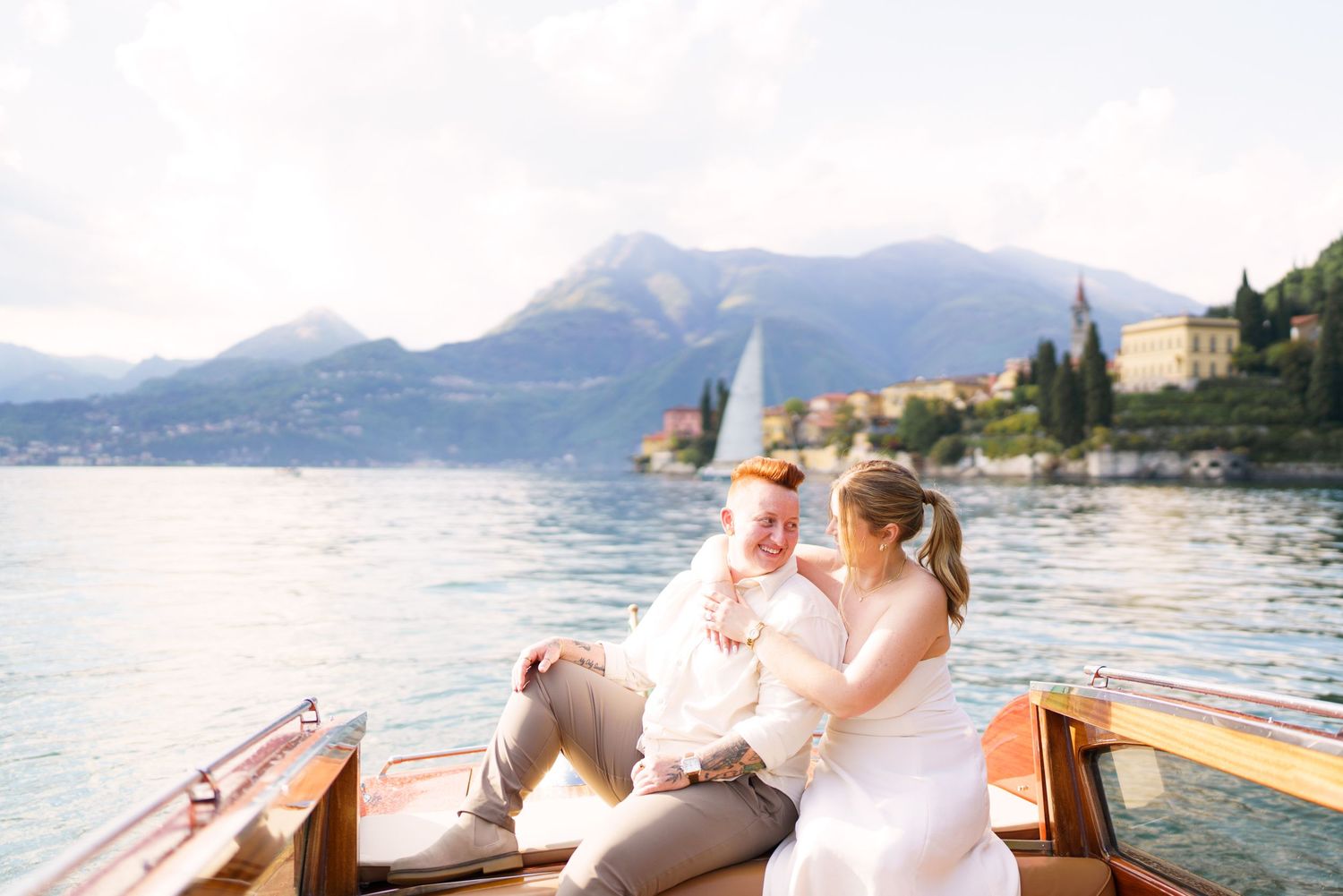 Couple in wedding attire sitting on a wooden boat on Lake Como, Italy, with mountain village backdrop.