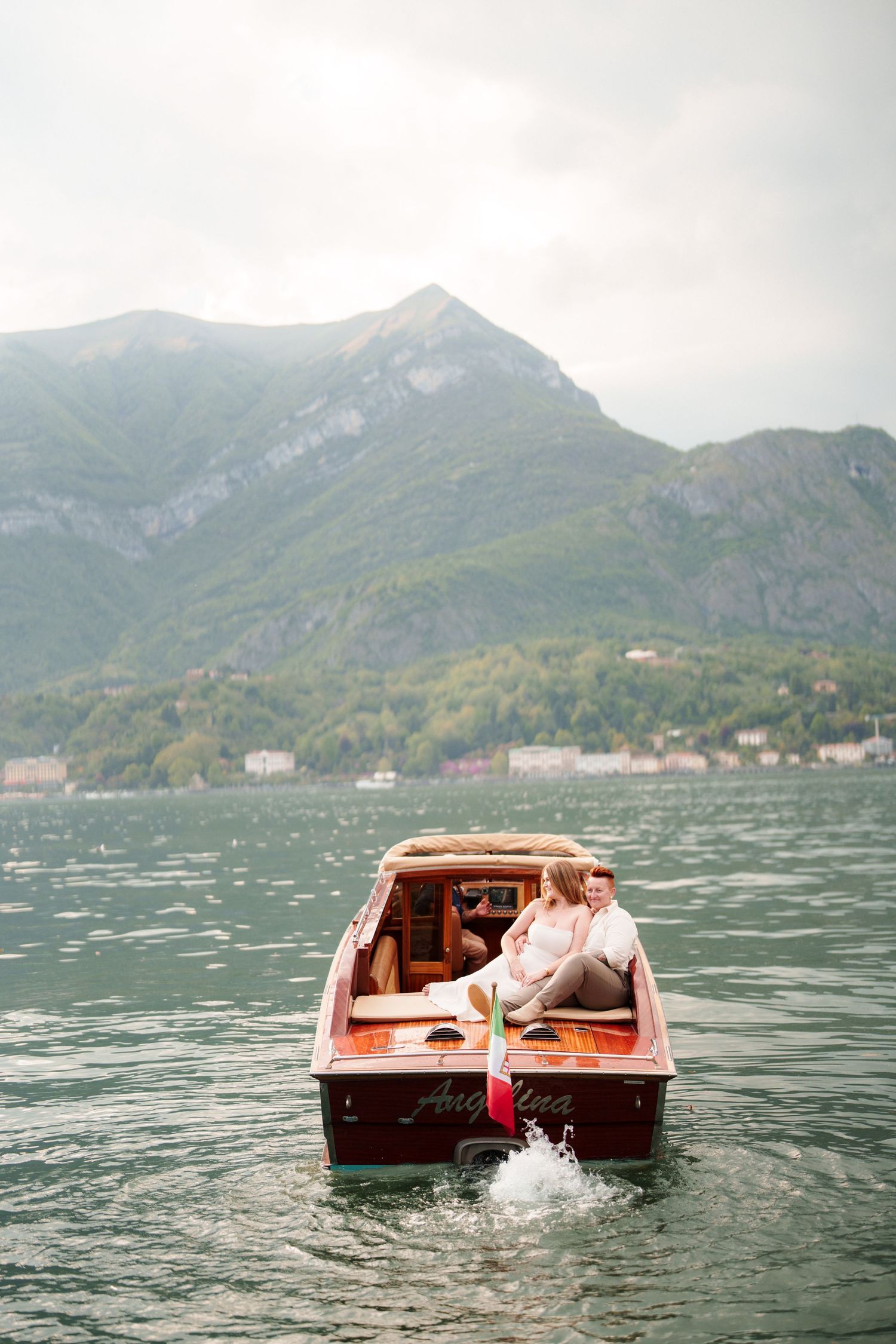 Couple riding a classic wooden boat on a scenic mountain lake under overcast skies.