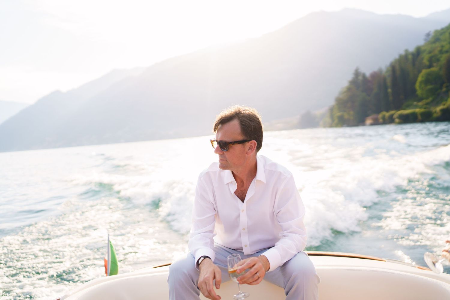 Man in white shirt and sunglasses relaxing on a luxury speedboat on a scenic mountain lake.