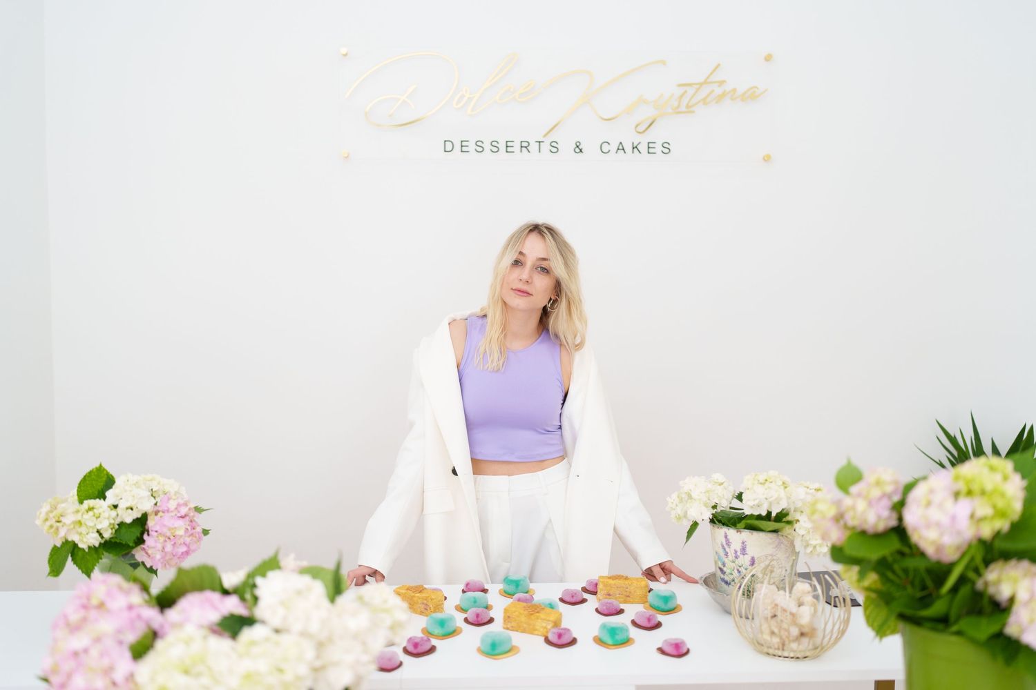 Woman in white blazer and lavender top poses at dessert table with macarons and flowers at Chelsea Krystina Desserts.
