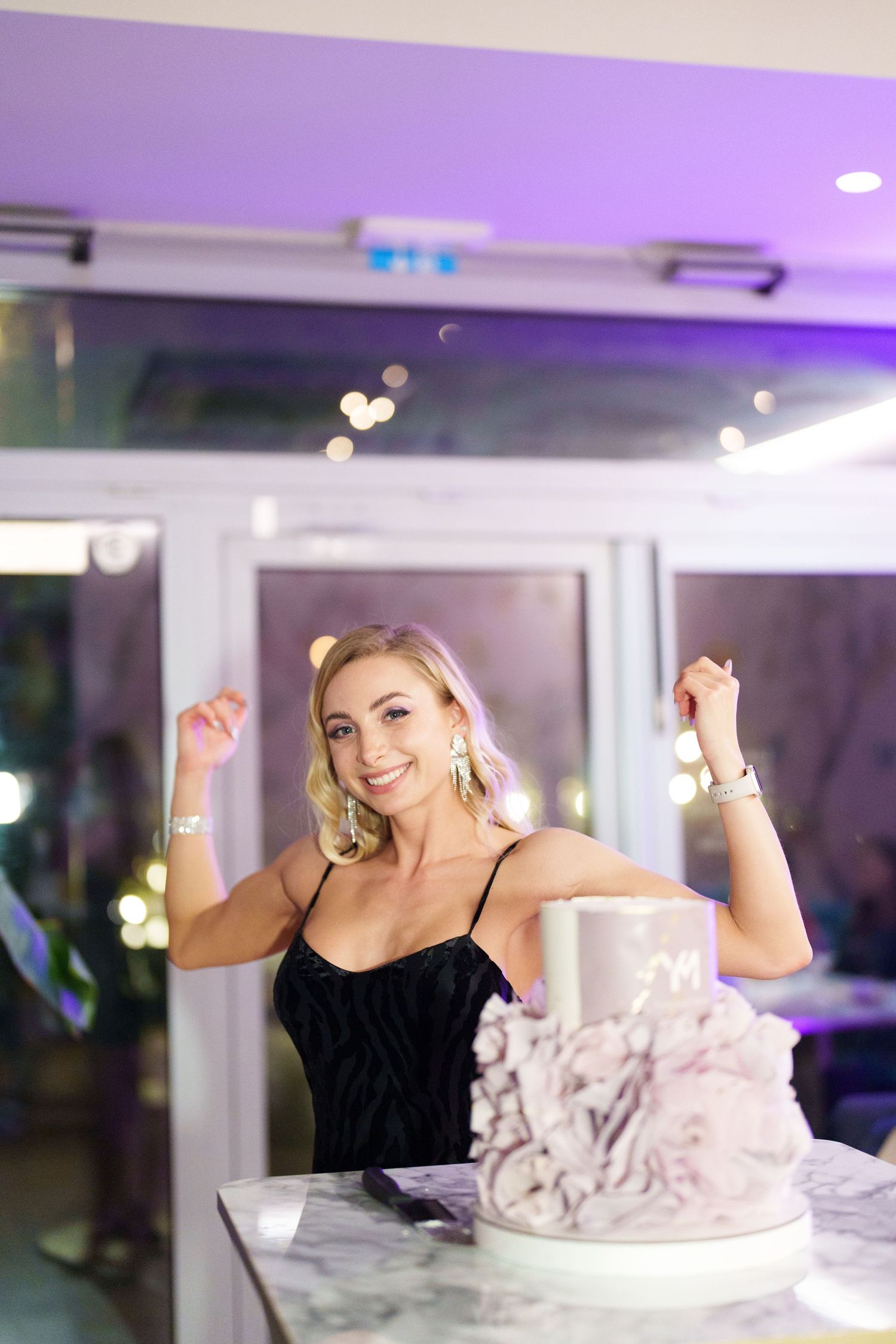 A smiling blonde woman in a black dress celebrates with raised fists beside a elegant white layered cake at a party.