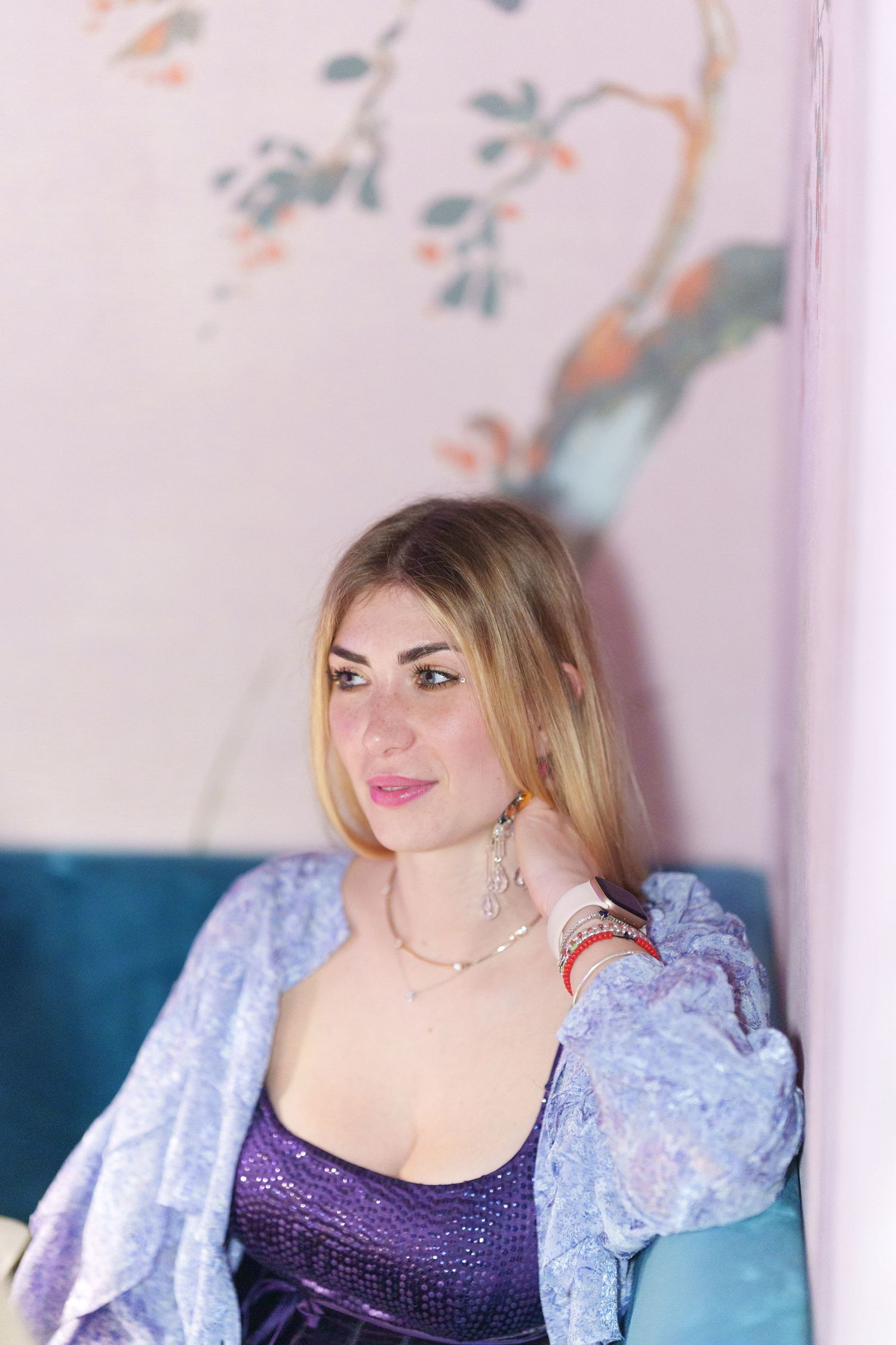 Blonde woman in purple sequin top and lace jacket poses on teal sofa before floral wall art.