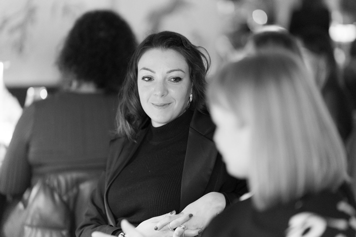 Black and white photo of a smiling woman in a black turtleneck engaged in conversation at a social event.