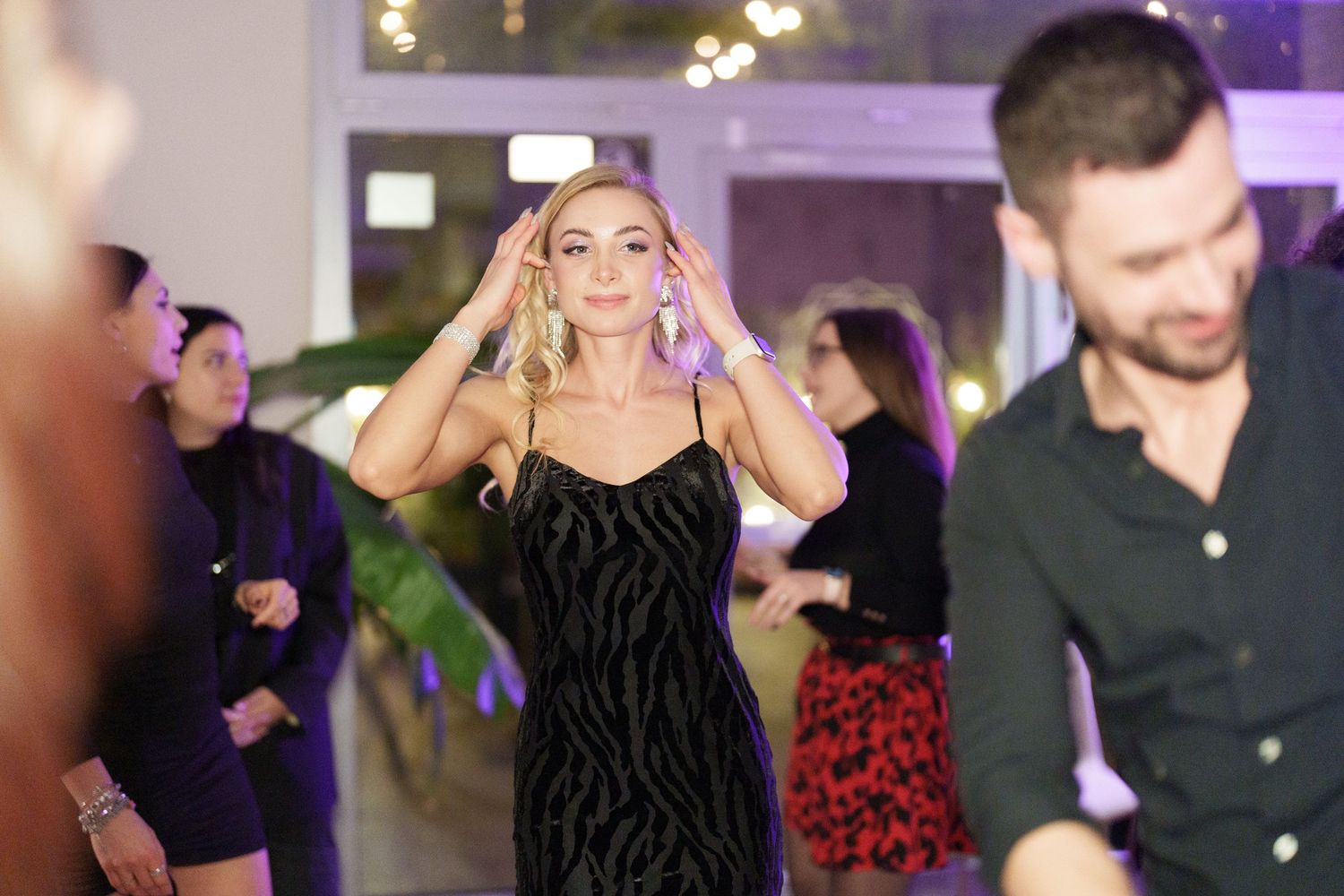 Woman in black velvet dress dancing at a party with guests socializing in the background.