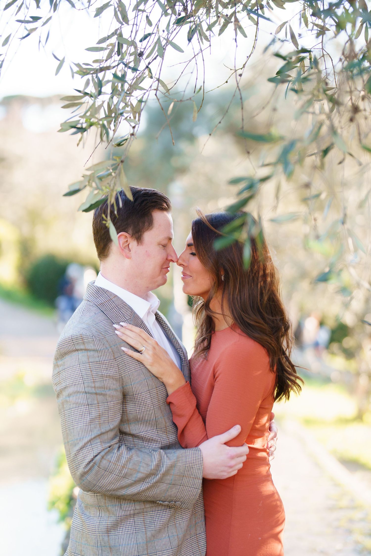 Couple sharing a romantic moment under olive tree branches, man in gray suit and woman in rust dress embracing outdoors.