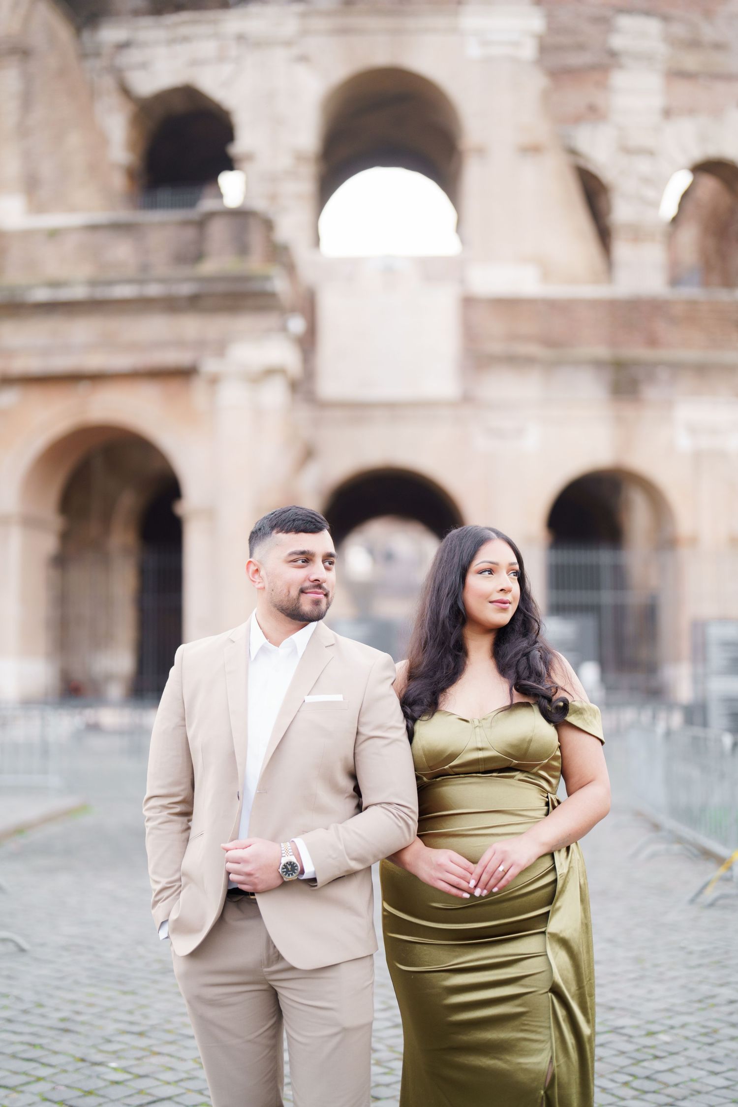 Elegant couple poses before the Colosseum in Rome, man in beige suit and woman in olive green gown.