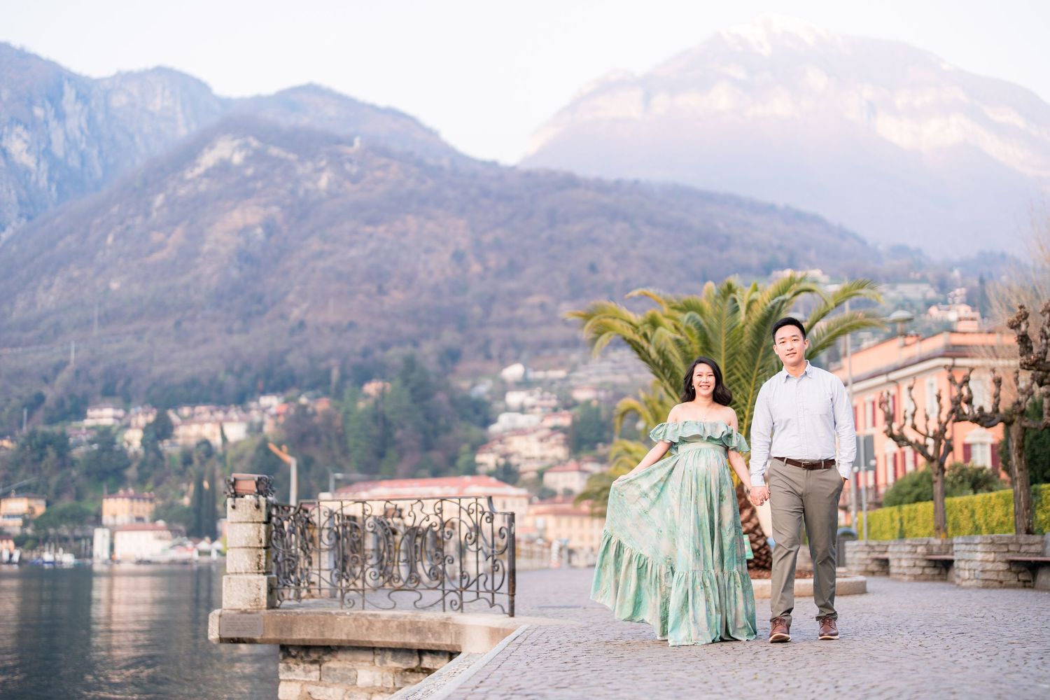 Couple walking hand-in-hand by Lake Como with mountains and village backdrop, woman in elegant green gown.