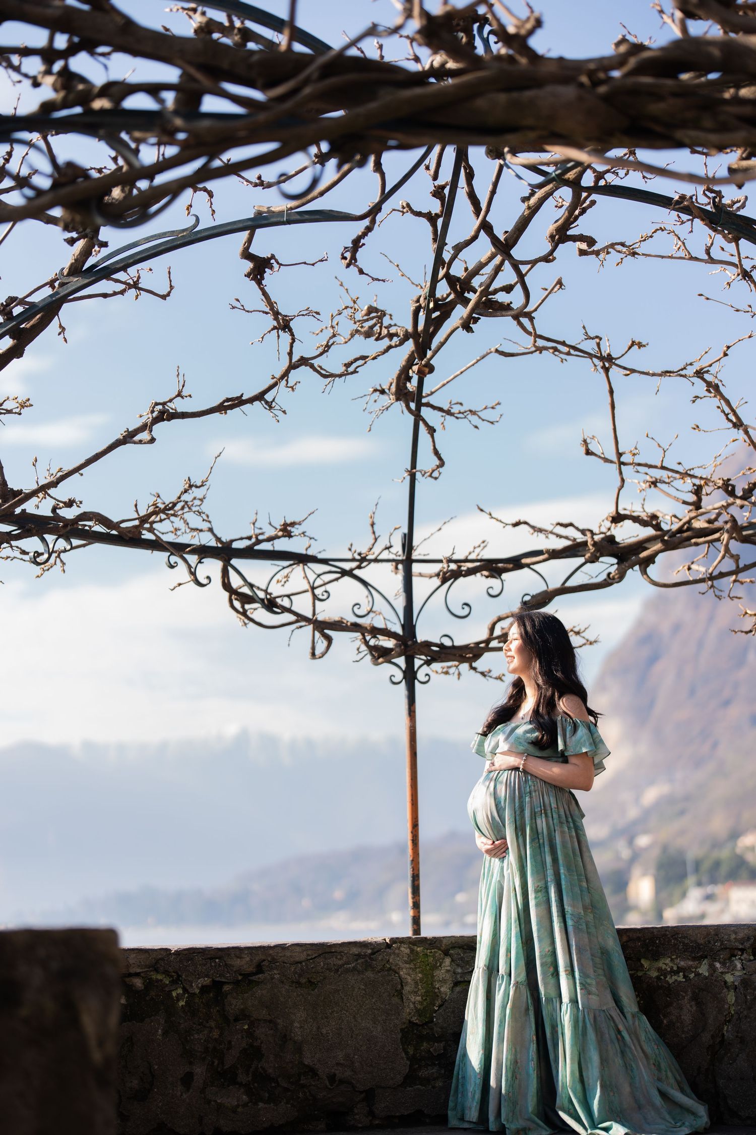 Pregnant woman in teal gown stands on stone terrace under bare vines with mountain backdrop.