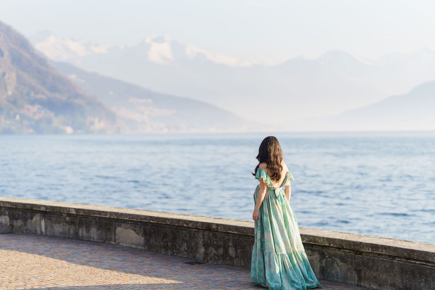 A woman in a flowing teal gown stands on a lakeside promenade with misty mountains in the background.