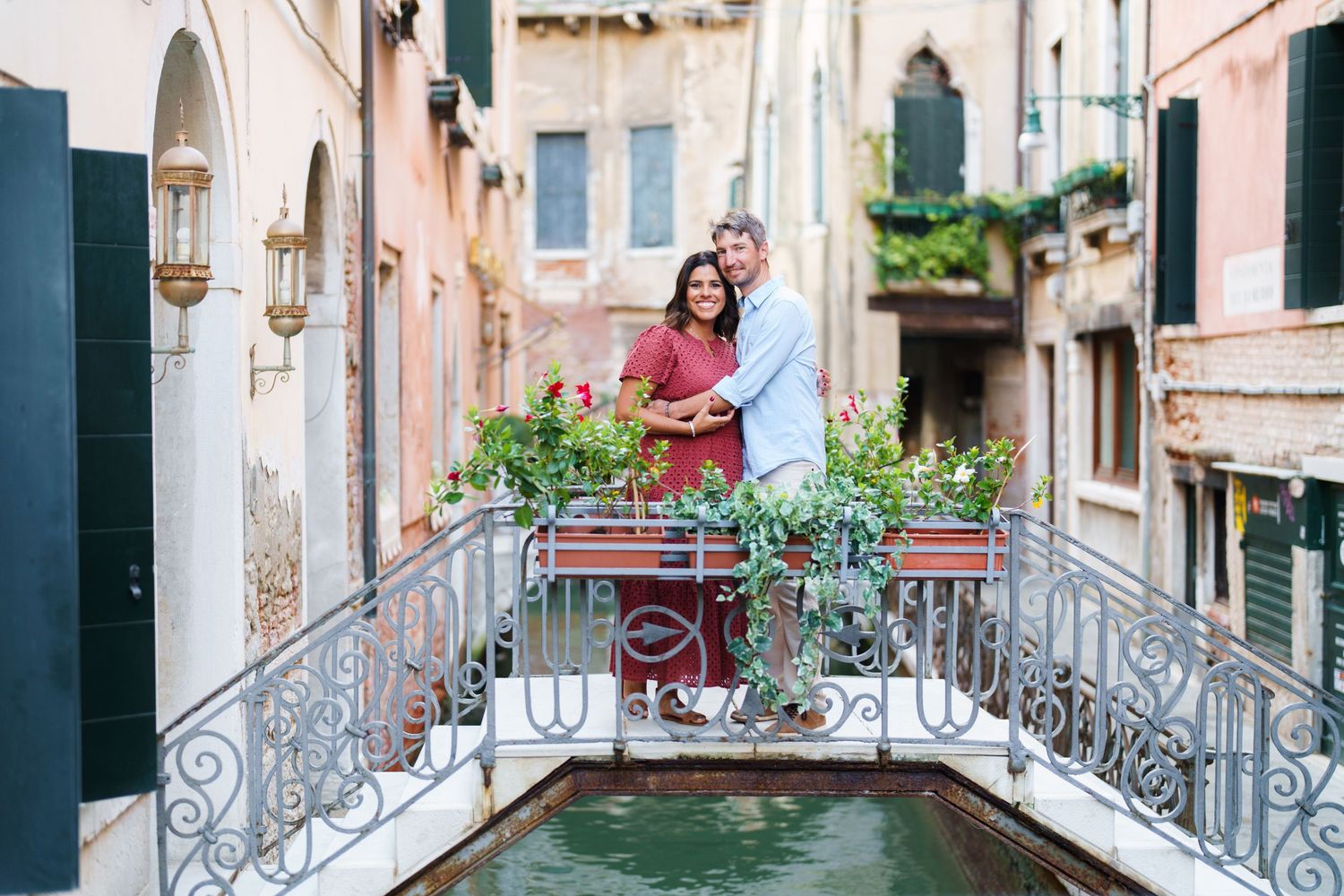Couple embracing on a romantic bridge over a canal in Venice, Italy, surrounded by colorful flowers and historic buildings.