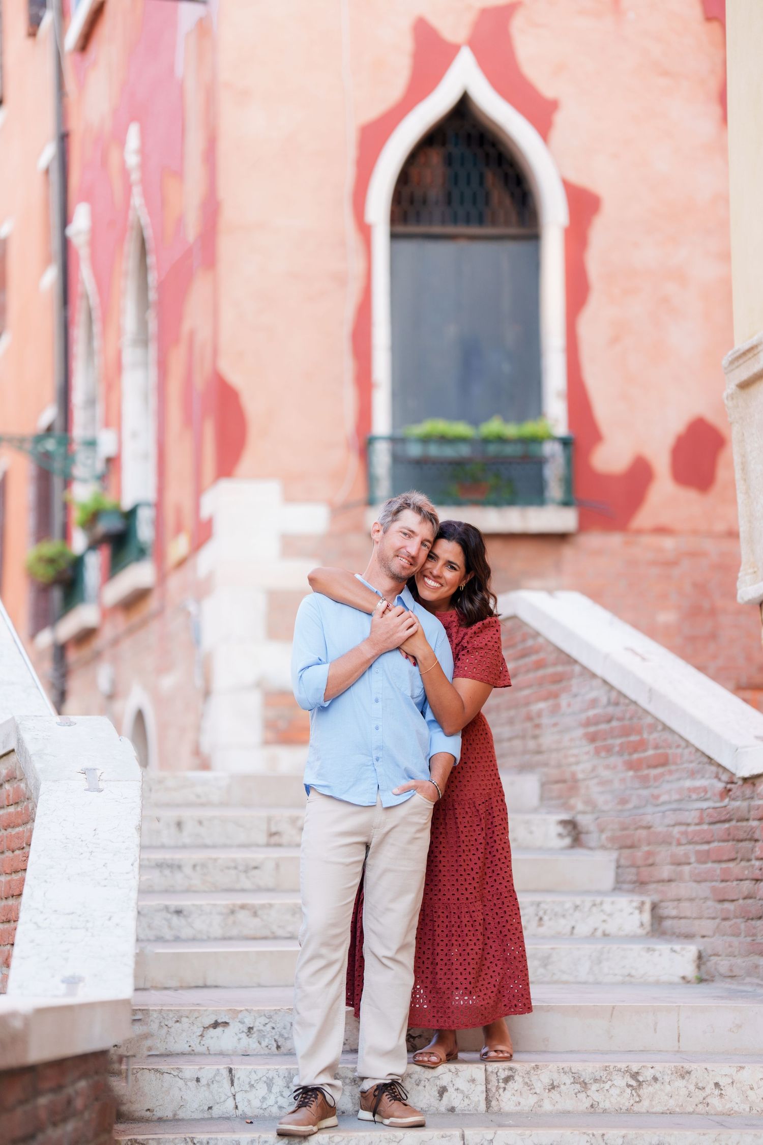 A couple embraces on stone steps in Venice, Italy, with a colorful red building and arched window behind them.