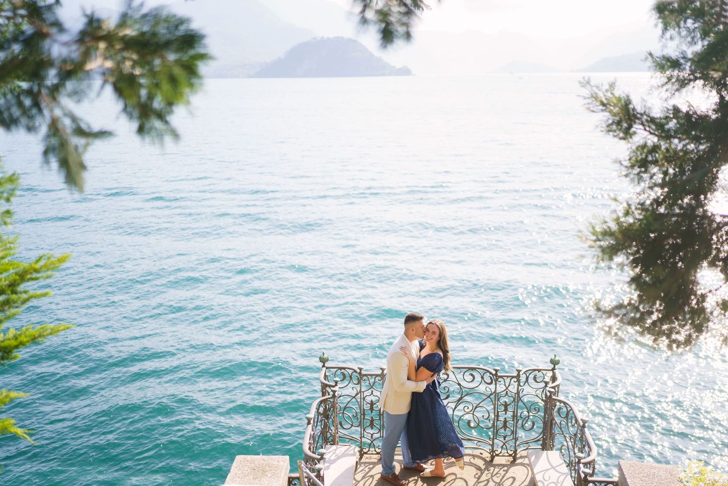 Romantic couple sits on ornate metal bench overlooking turquoise ocean waters on sunny tropical coastline.