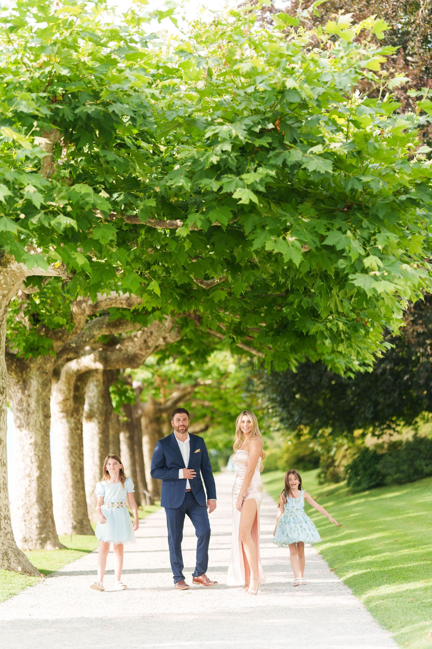 Family walking together along a tree-lined path in a lush green park on a sunny summer day.