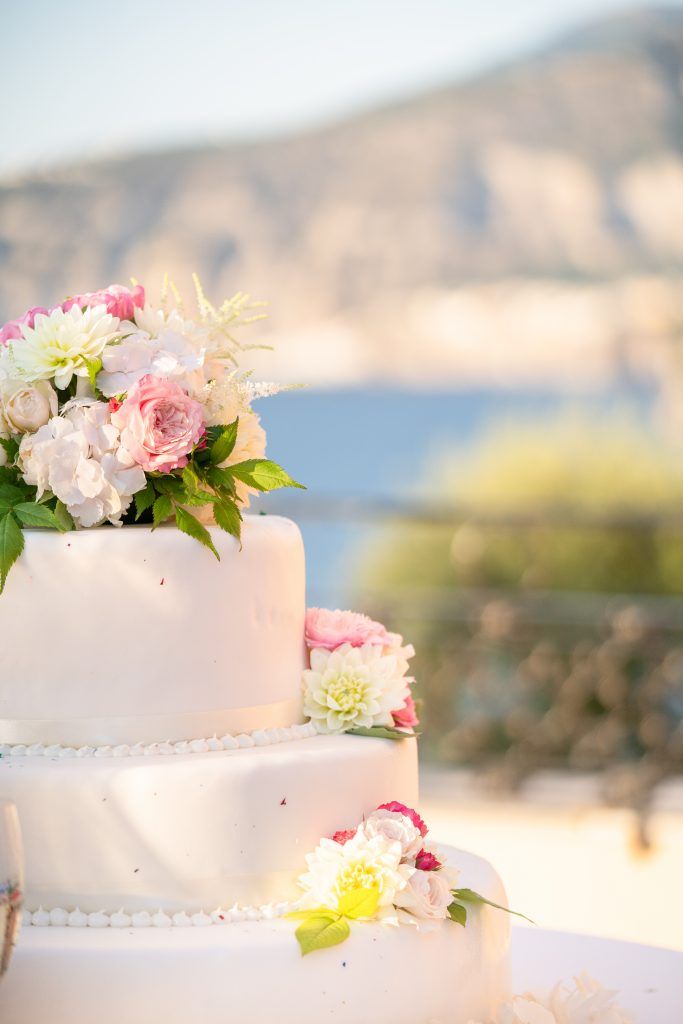 Elegant white wedding cake decorated with fresh pink flowers against a coastal backdrop.