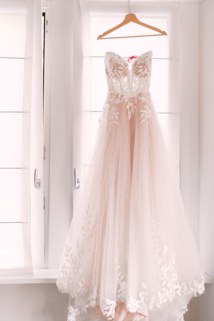 Blush wedding gown with lace details hanging in bright white room.