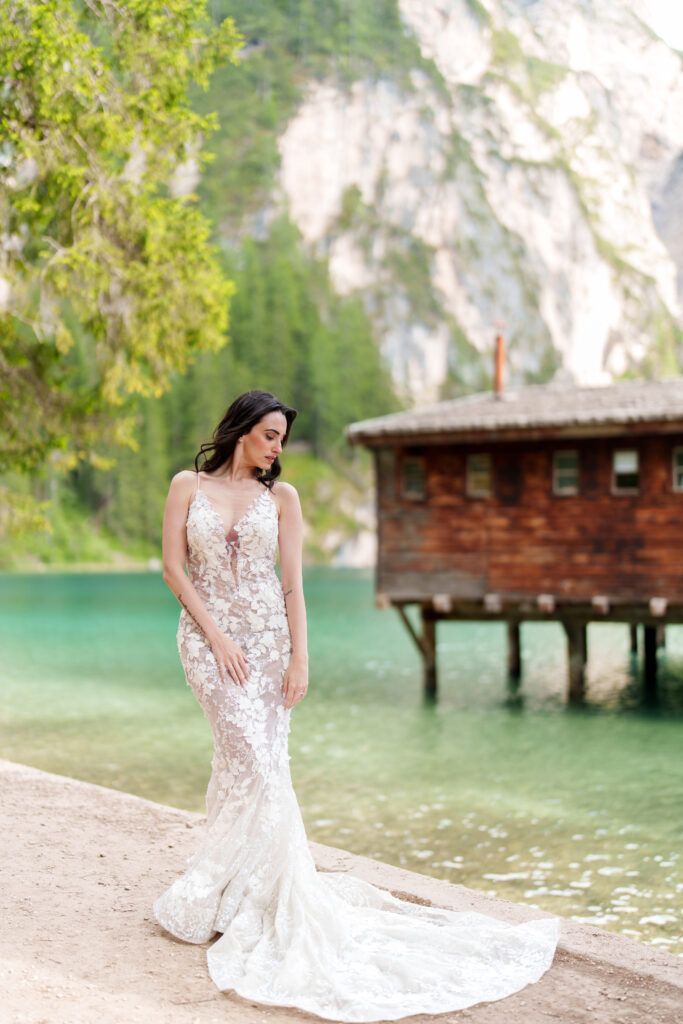 A bride in a lace wedding dress poses near a rustic wooden boathouse on a scenic mountain lake.