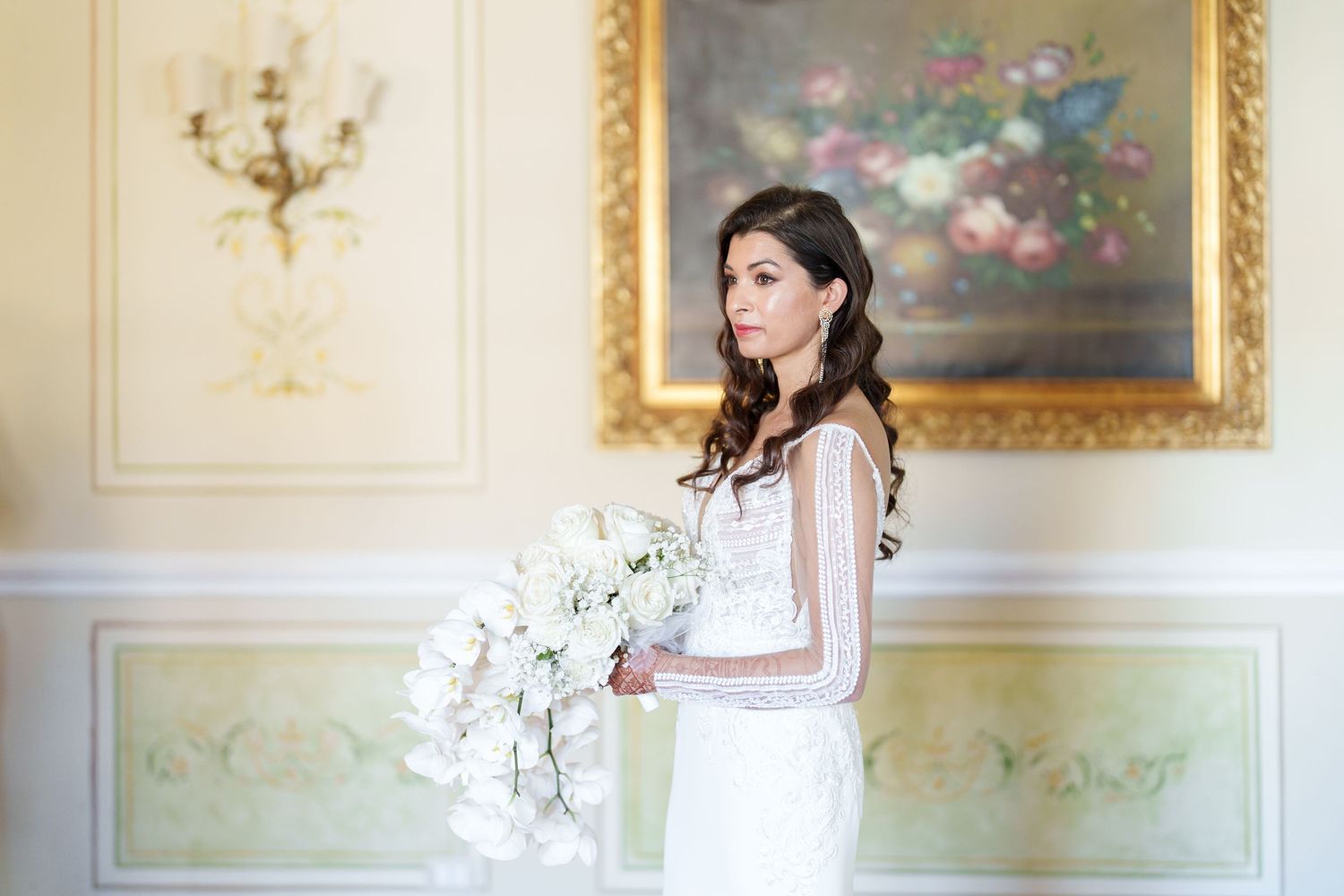 Women in white dresses pose elegantly on a green velvet sofa in an ornate room with floral artwork.