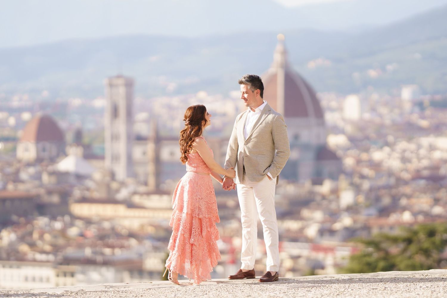A couple in formal attire stands at a scenic overlook with Florence's Duomo cathedral and cityscape in the background.