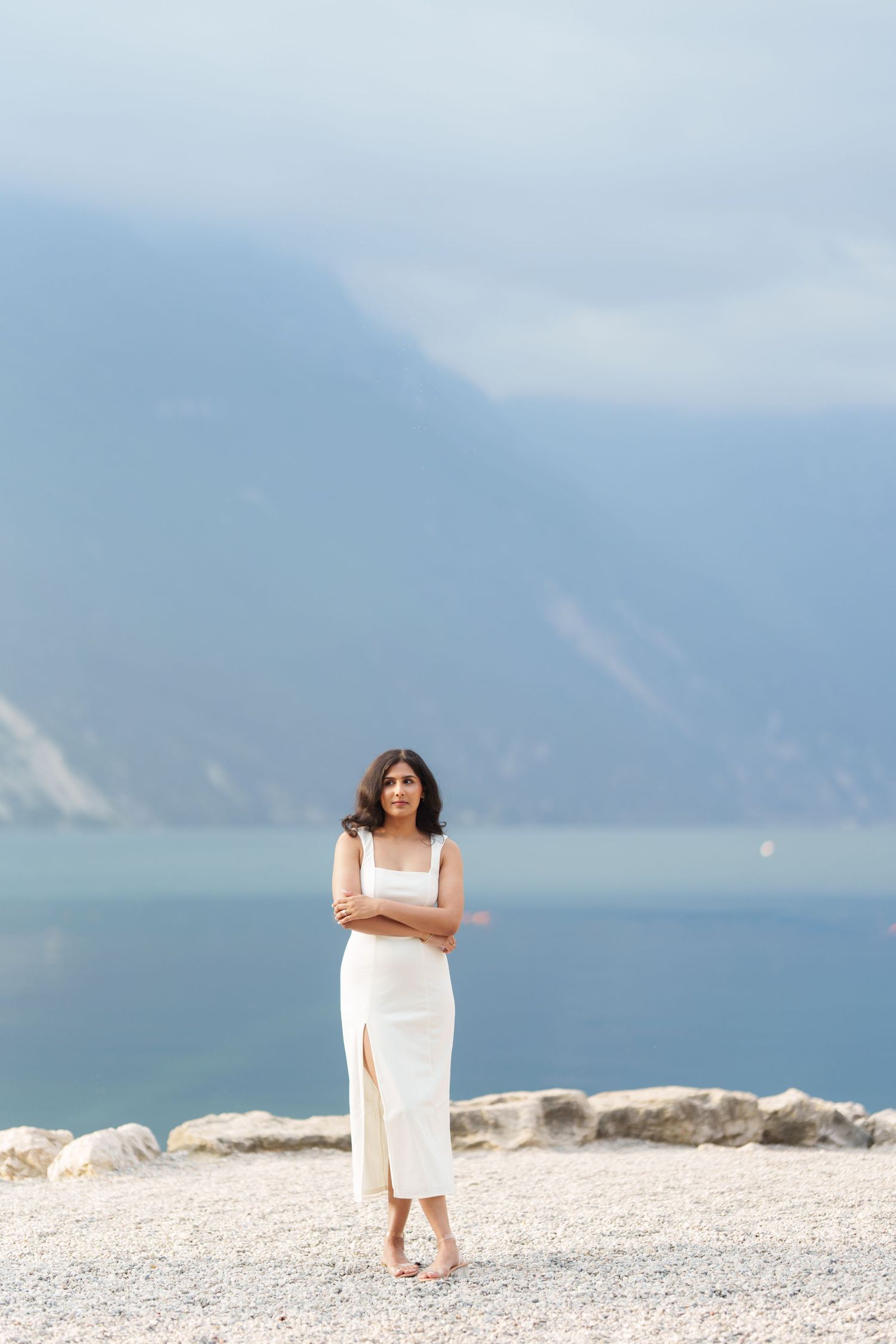 A figure in a white dress stands on a rocky beach with misty mountains and water in the background.