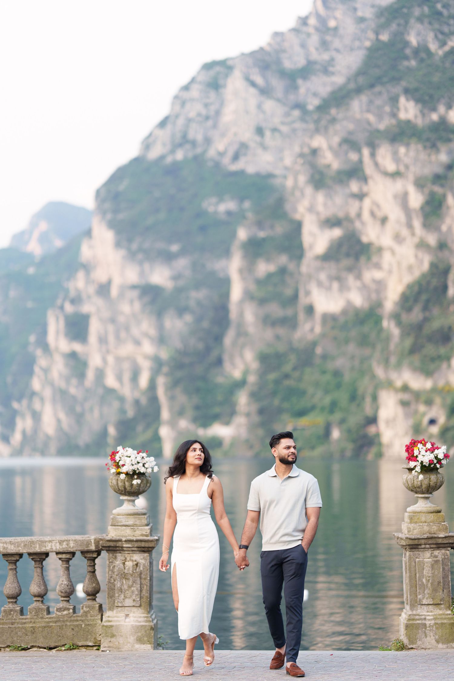 A couple strolls hand in hand along a stone balustrade by a scenic mountain lake with dramatic cliffs in the background.