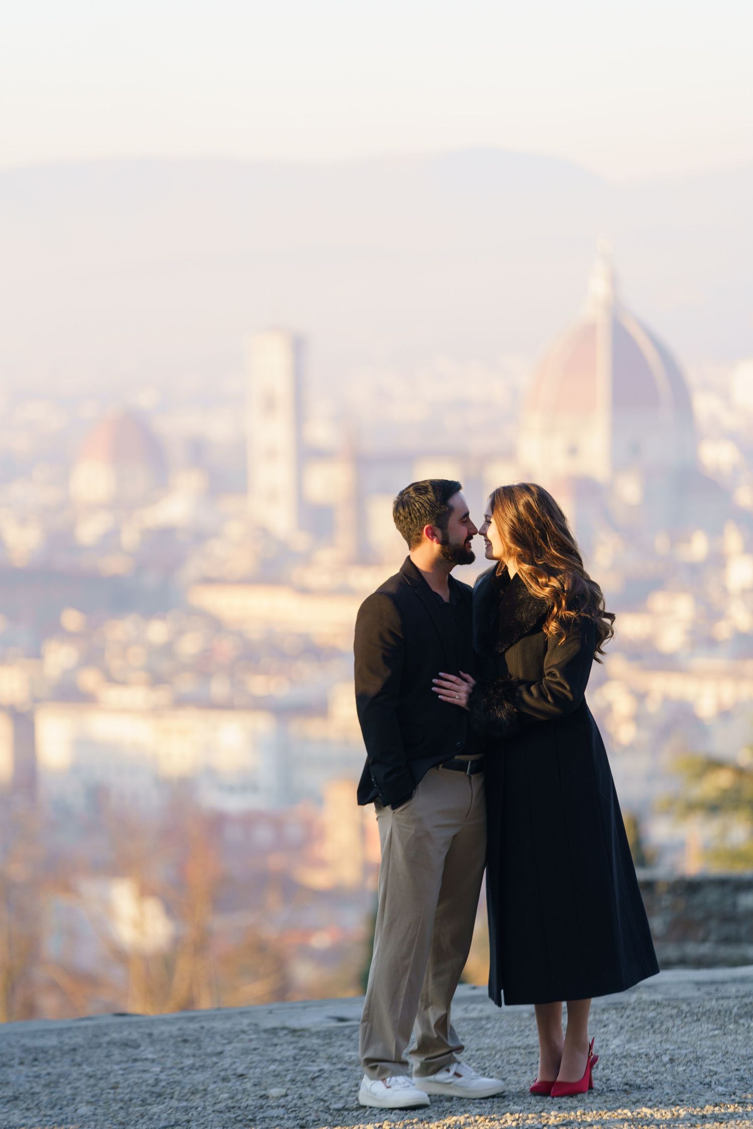 A couple sits together on a hilltop overlooking a misty cityscape during sunset.