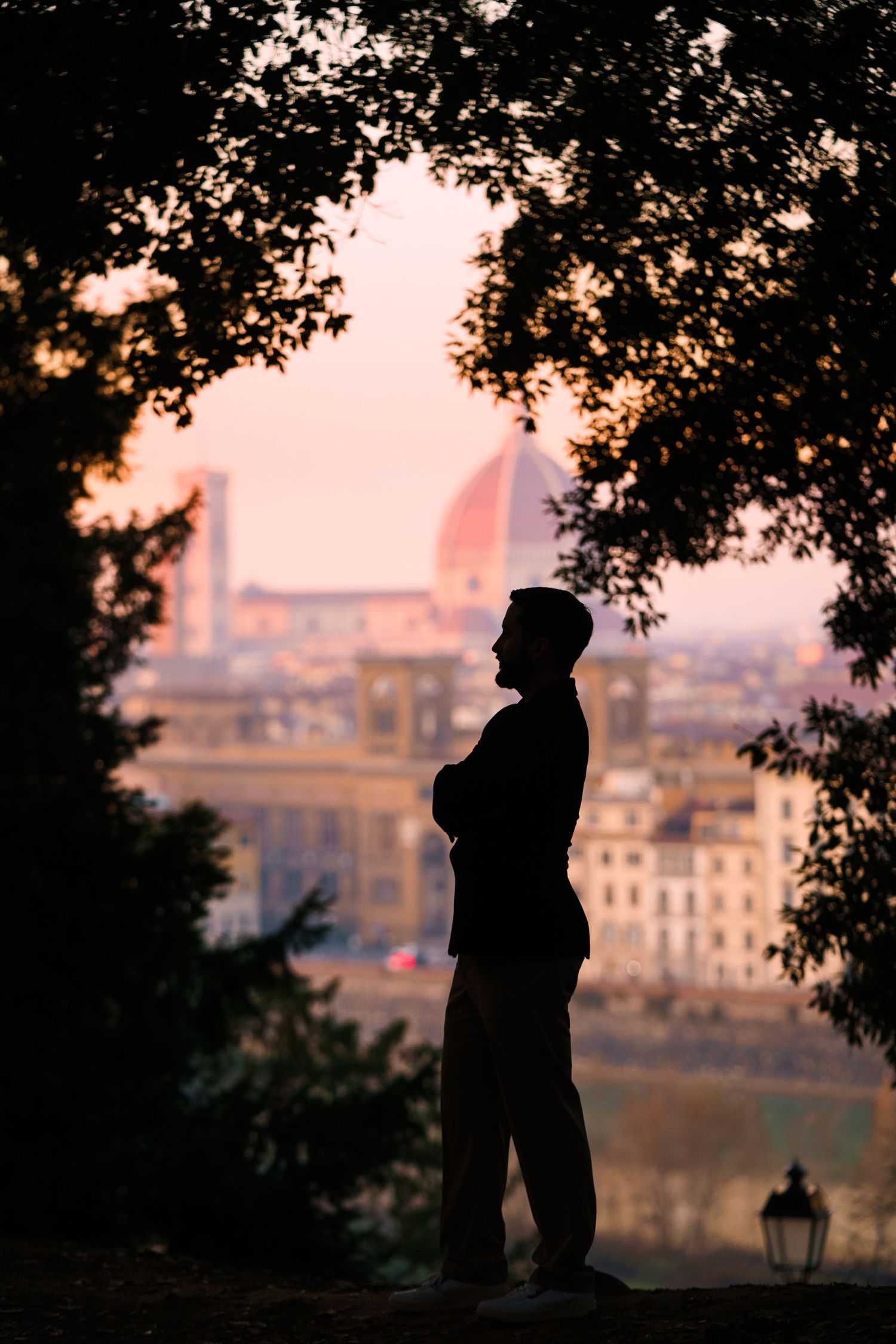Silhouette of a figure standing against a sunset cityscape of Florence, Italy with trees framing the scene.