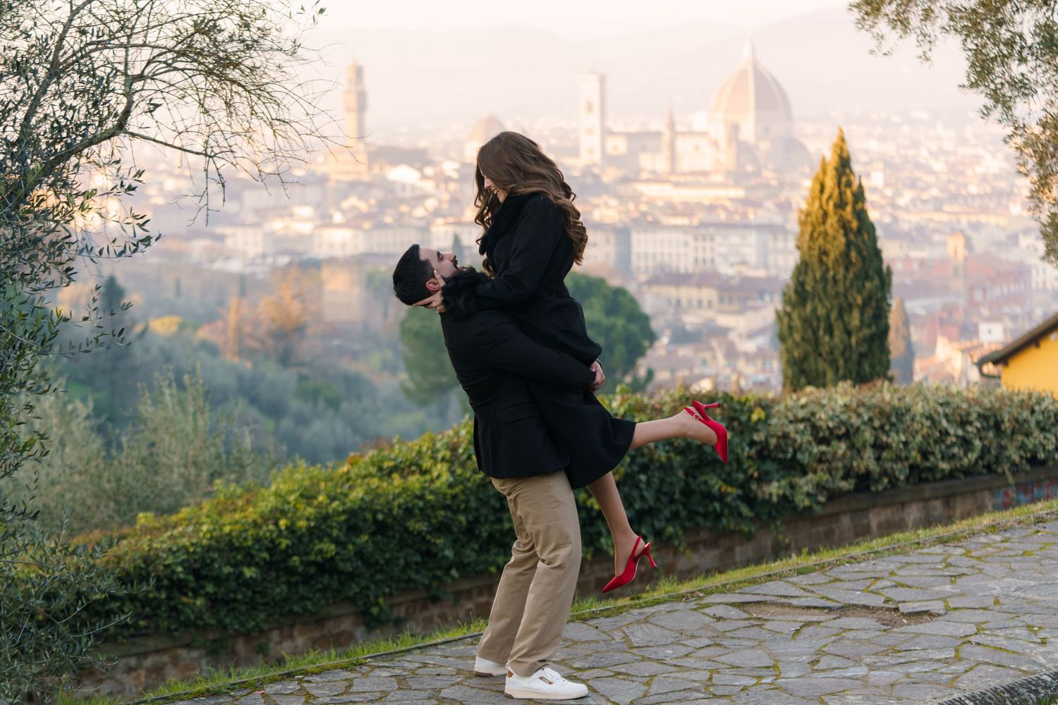 A couple shares a romantic moment on a hilltop path overlooking a city vista at dusk.